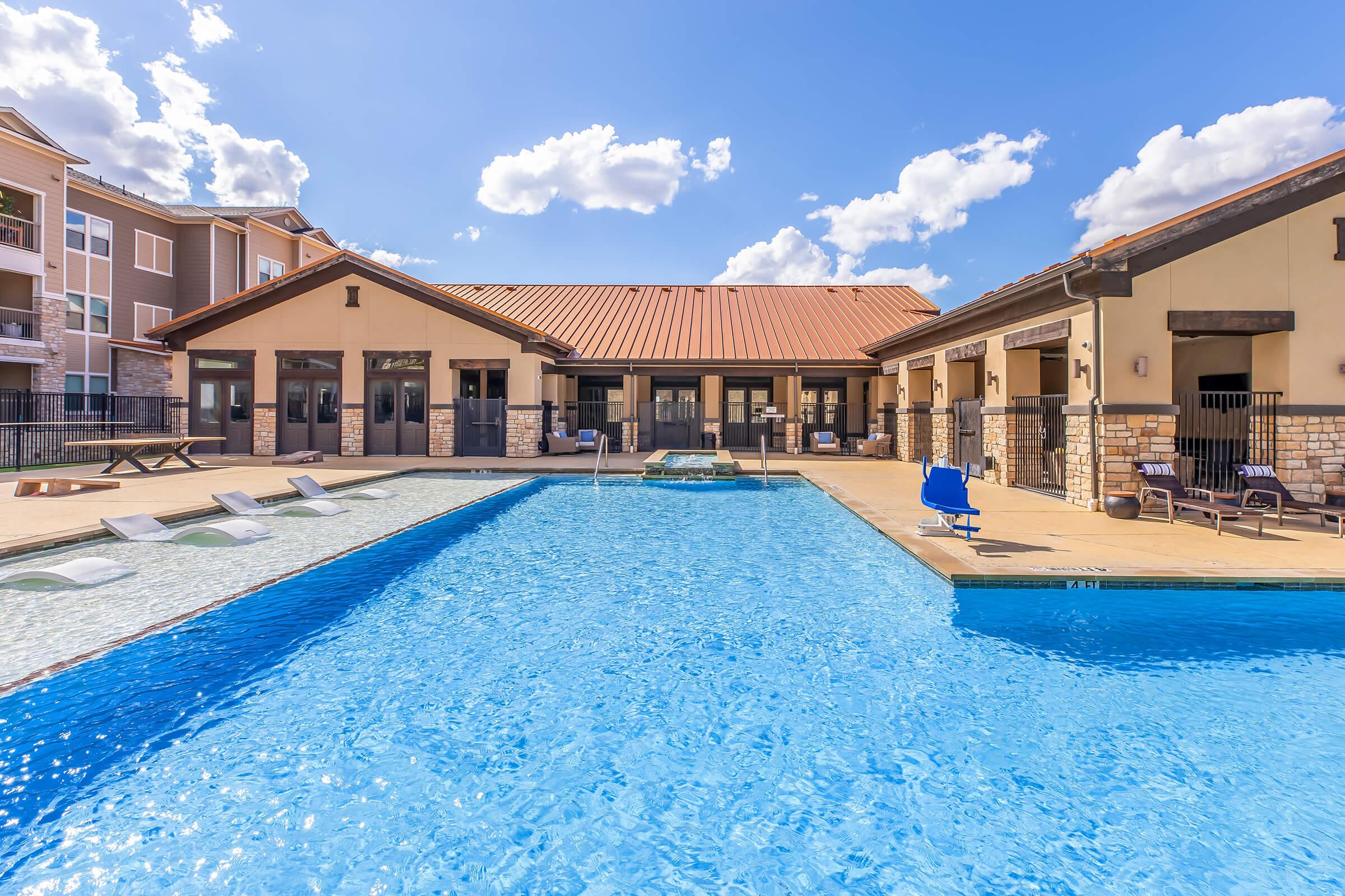 A large outdoor swimming pool with clear blue water, surrounded by lounge chairs. In the background, there's a clubhouse with a reddish-brown roof and multiple windows. The sky is bright with fluffy white clouds. Sunlight reflects off the water, creating a inviting atmosphere for relaxation and recreation.