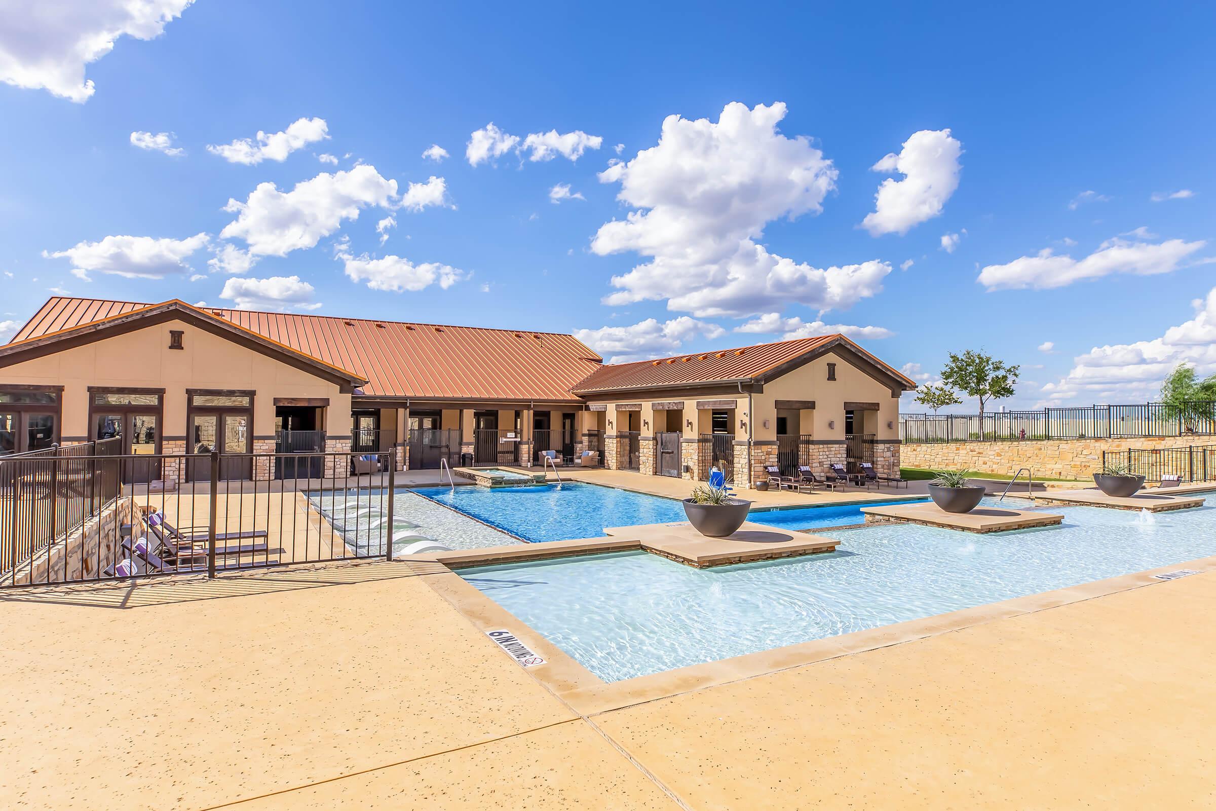 A sunlit outdoor area featuring a swimming pool with lounge chairs, surrounded by a smooth concrete deck. The backdrop includes a modern building with a red tile roof and a clear blue sky dotted with fluffy white clouds. Potted plants are placed near the pool, enhancing the tranquil ambiance.