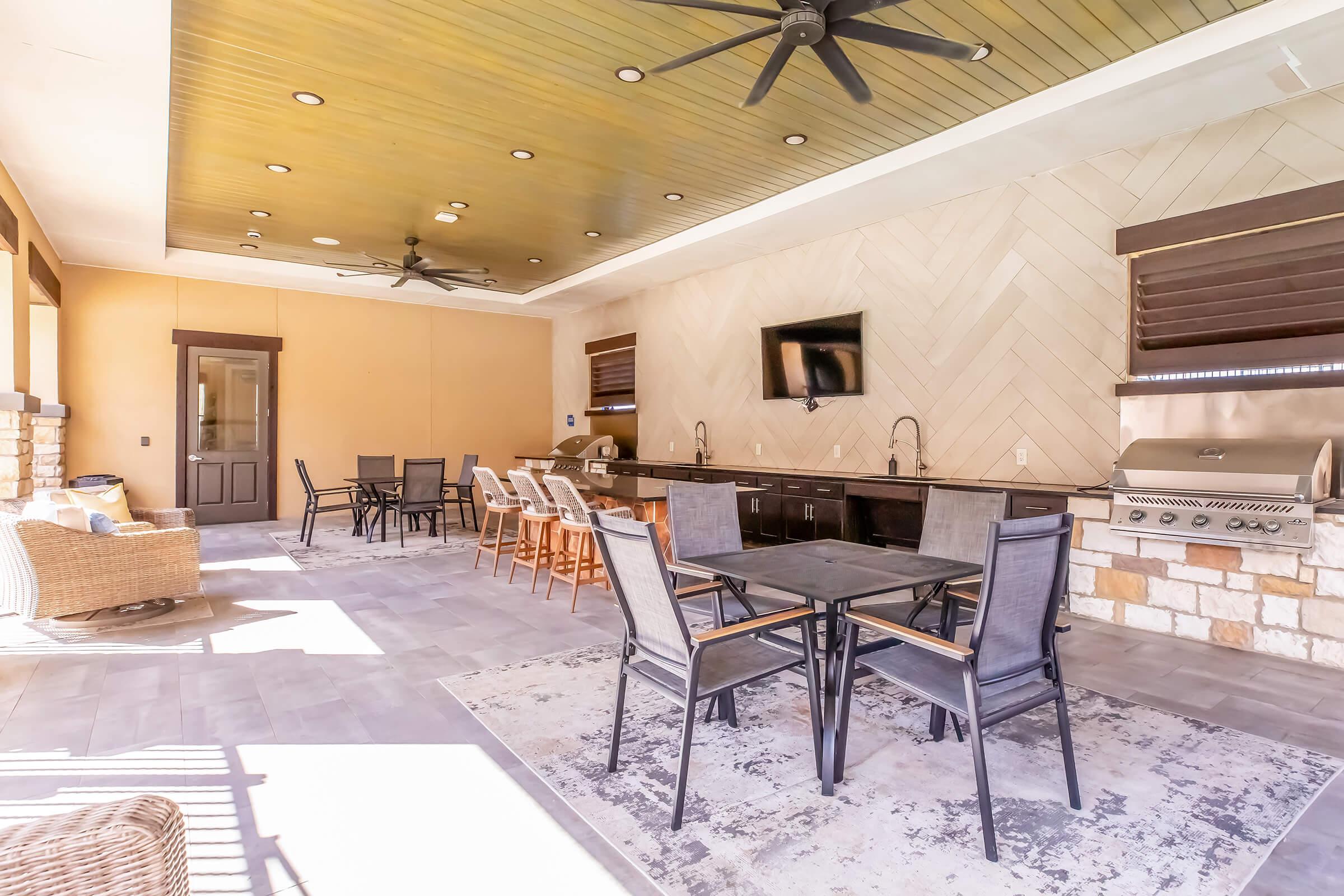 Large outdoor living space featuring comfortable seating, a dining area, and a kitchen setup with a barbecue grill. The ceiling has wooden panels and modern fans, while the stone accents add an elegant touch. Natural light fills the area through large windows.