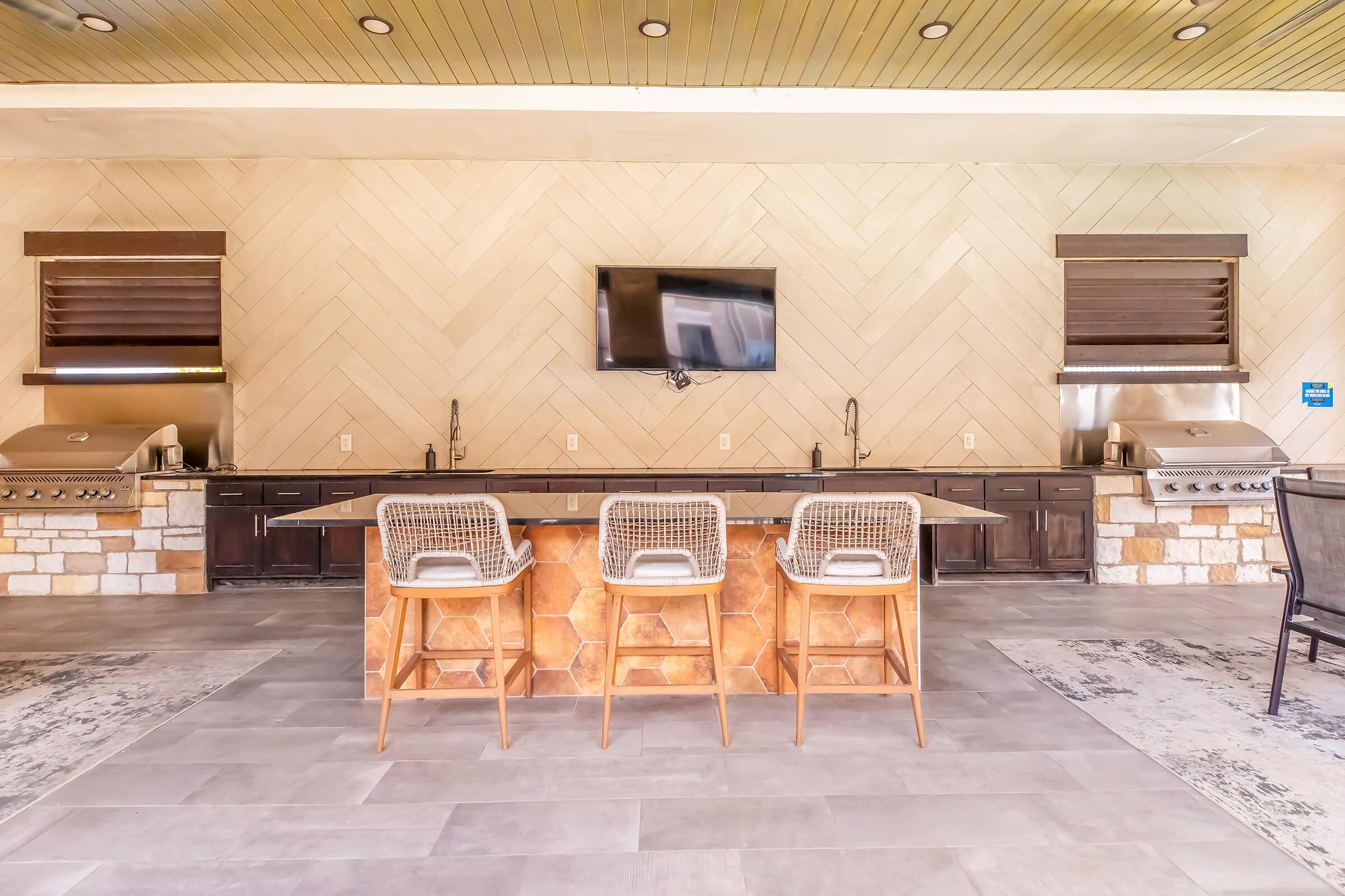Outdoor kitchen area featuring a long countertop with three bar stools, two stainless steel grills, and a wall-mounted television. The space has a light-colored tiled wall and a wooden ceiling, creating a modern and inviting atmosphere.