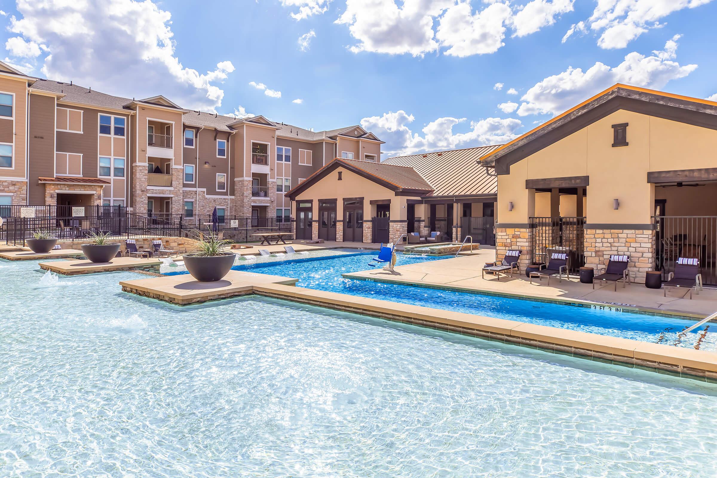 A serene outdoor pool area with clear water, lounge chairs, and shaded cabanas. Surrounding the pool are modern apartment buildings with a contemporary design, under a bright blue sky dotted with fluffy white clouds. The area looks inviting and well-maintained, ideal for relaxation and leisure.