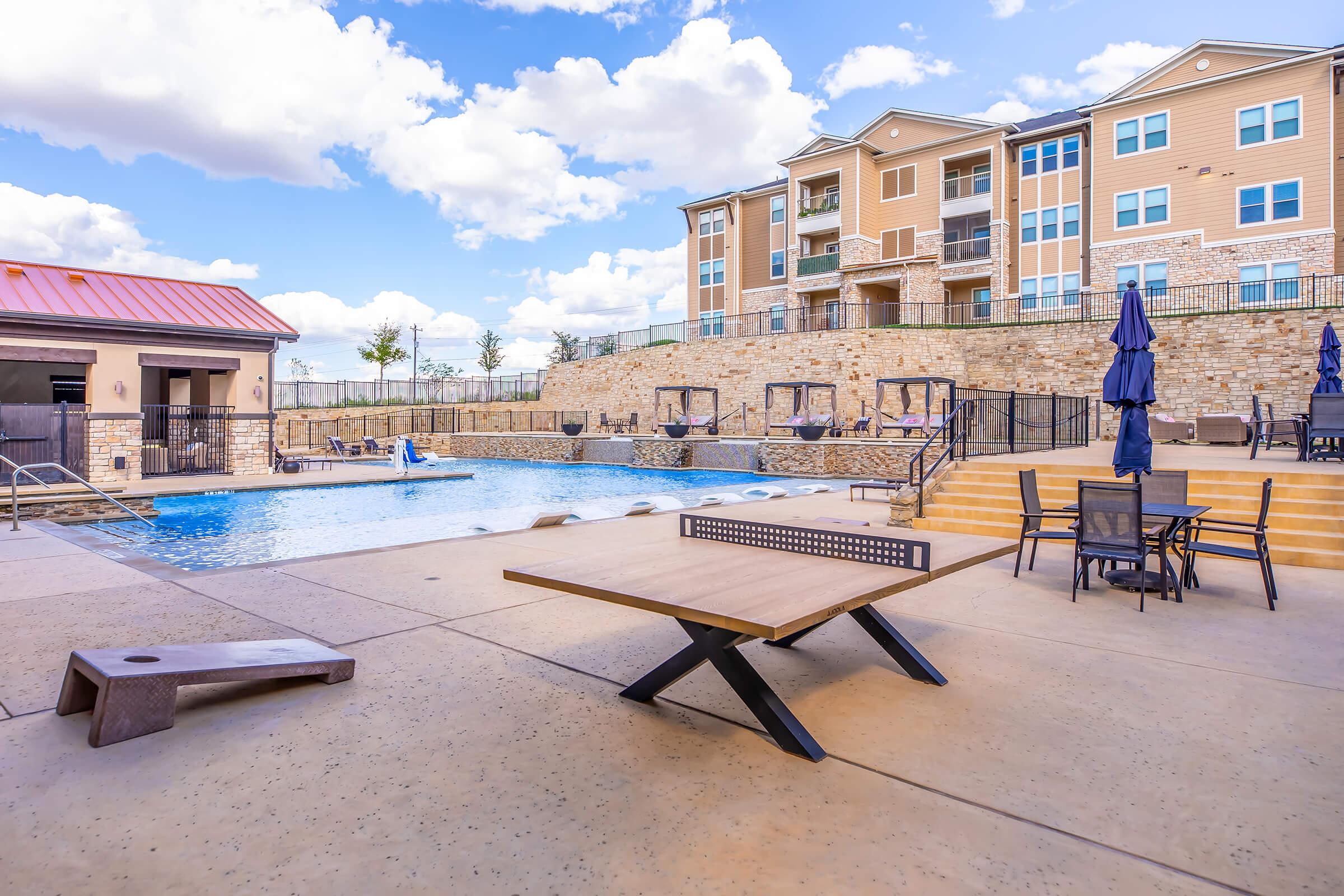 A spacious outdoor pool area featuring a large swimming pool, lounge chairs, and shaded umbrellas. Surrounding the pool are stone structures and an apartment building in the background, with blue skies and fluffy clouds overhead. A modern patio table and chairs are positioned in the foreground.