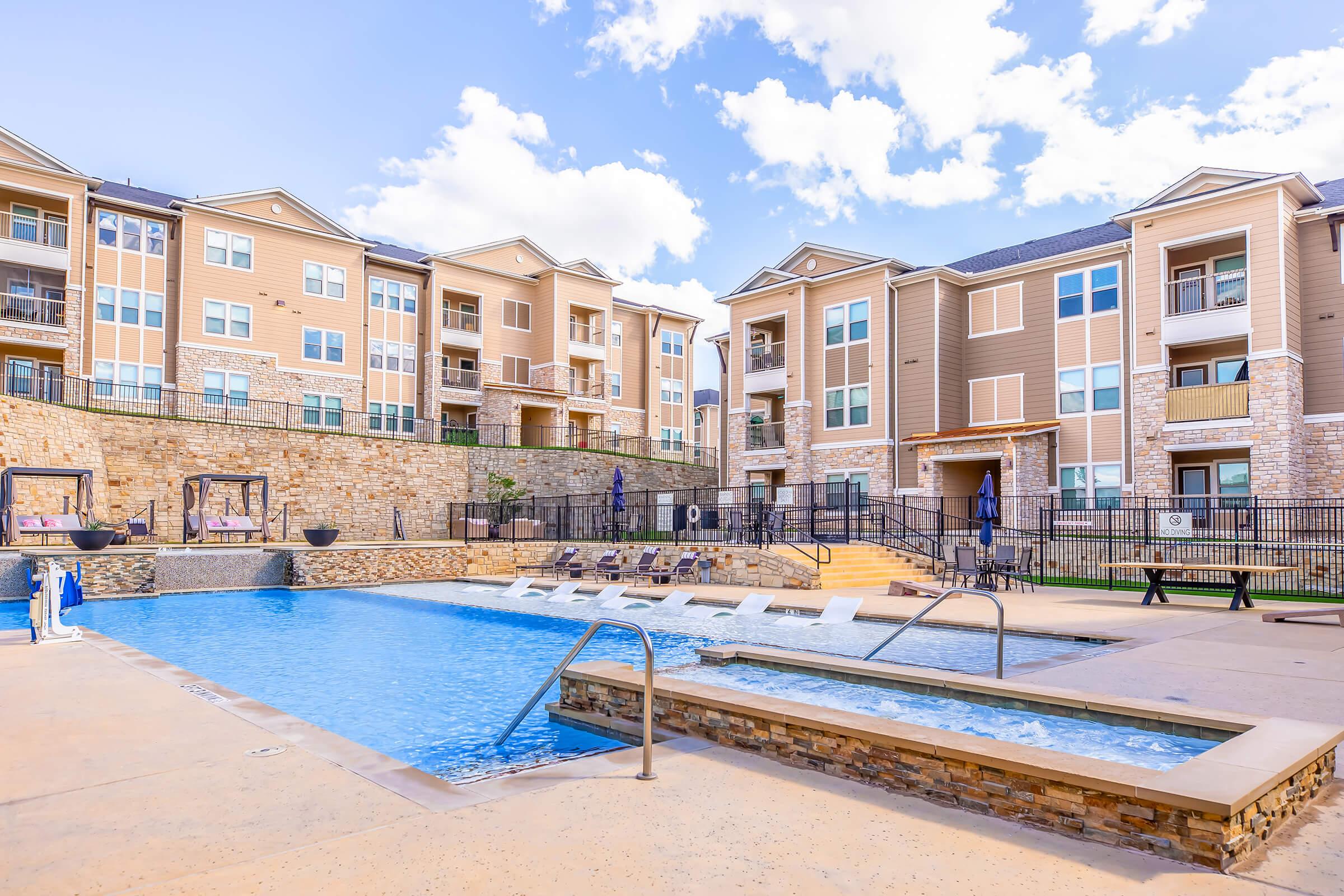 A swimming pool with lounge chairs surrounded by a landscaped area and modern apartment buildings. The pool area features a hot tub and is designed for relaxation, with blue skies and fluffy clouds above. The setting is inviting and well-maintained, ideal for outdoor leisure activities.