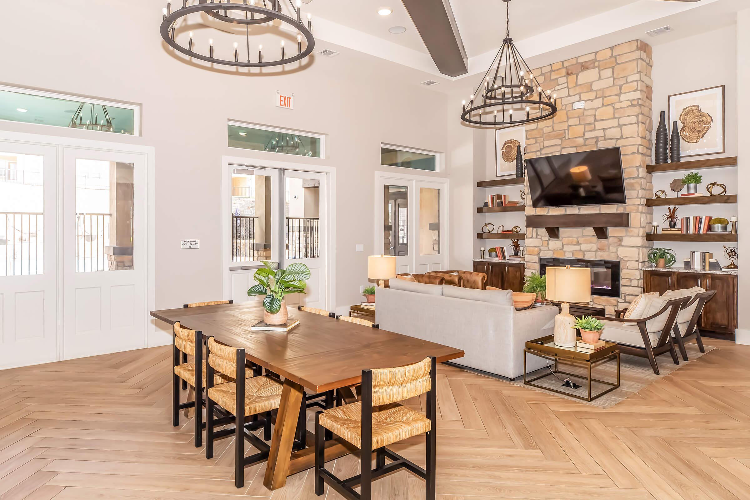 A spacious and stylish living area with a wooden dining table and chairs, a cozy sofa, and a stone fireplace. Large windows let in natural light, and decorative plants and artwork add a warm touch. Modern chandeliers hang from the ceiling, enhancing the inviting atmosphere.