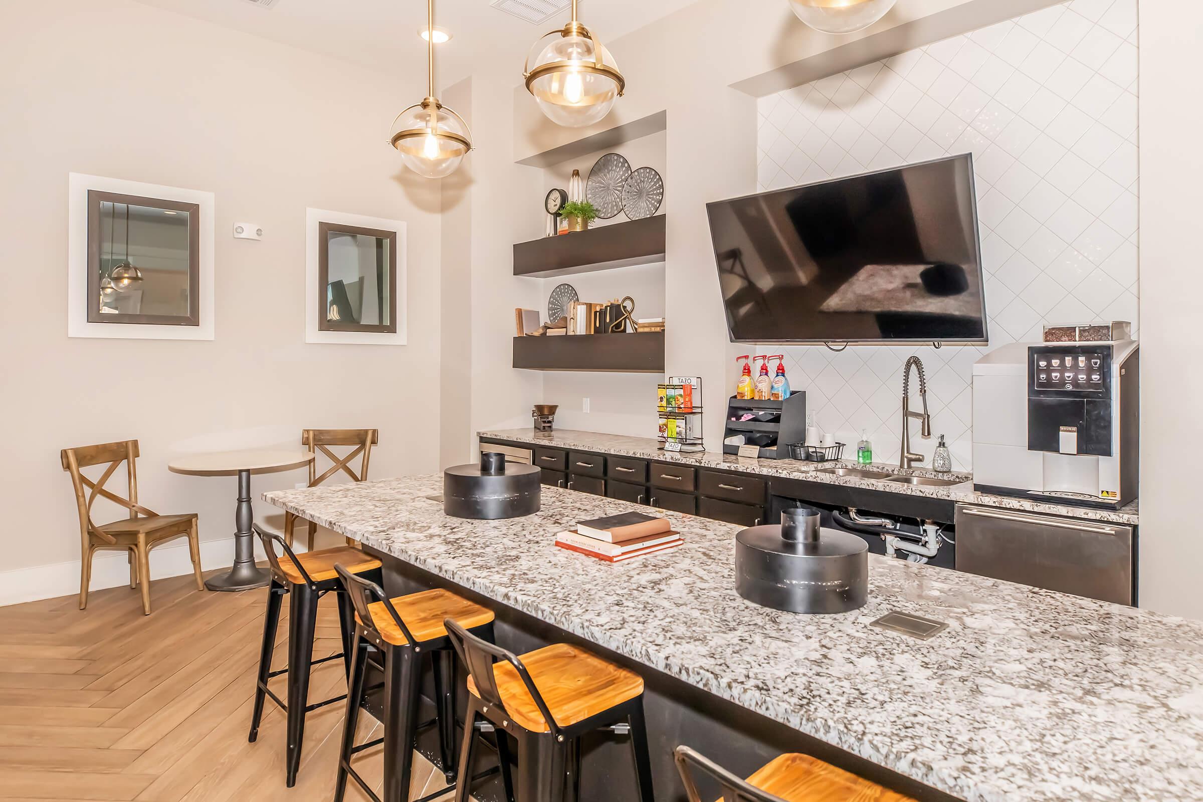 A modern kitchen or coffee bar featuring a gray granite countertop with seating, a TV mounted on the wall, shelves displaying decorative items, and a coffee machine. The space has warm lighting from pendant lamps and a small dining table with two chairs. The overall aesthetic is contemporary and inviting.