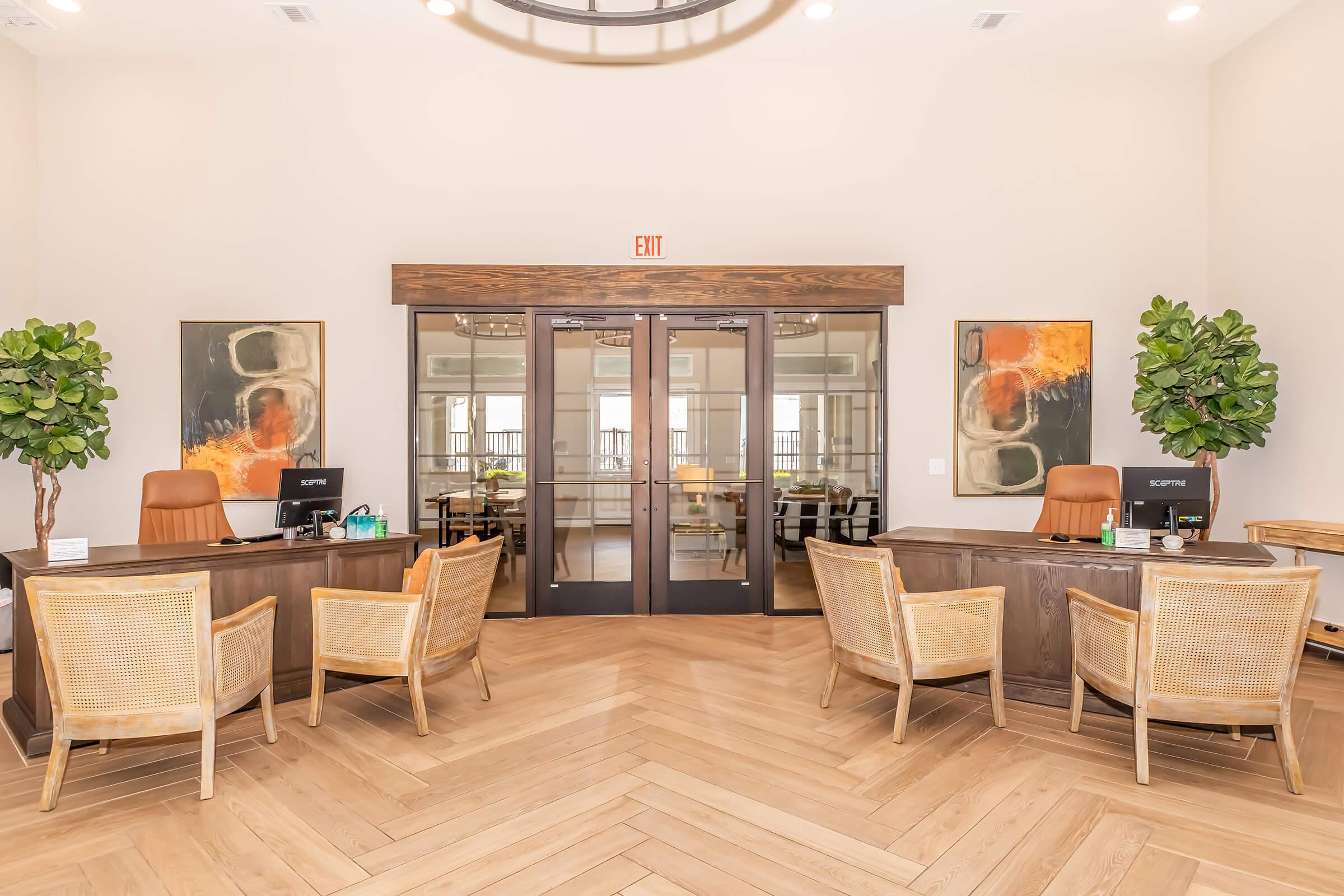 Bright and modern reception area featuring two desks with computers and comfortable chairs. Each desk is adorned with decorative items, and the space is enhanced by large artwork and potted plants. The flooring has a herringbone pattern, contributing to a welcoming atmosphere.