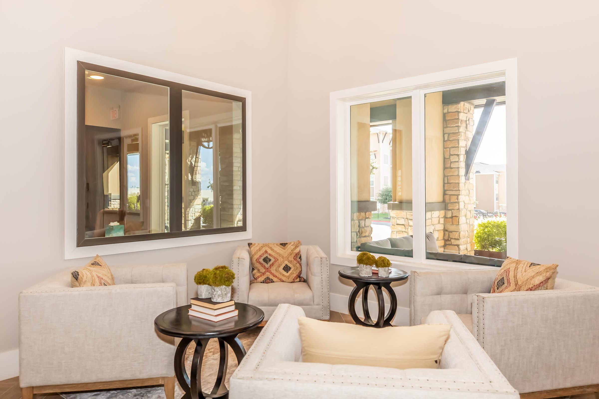 A cozy seating area with two light-colored sofas and decorative pillows, two round coffee tables in the center, and large windows letting in natural light. The walls are painted a soft color, and there are potted plants on the tables, creating a welcoming atmosphere.