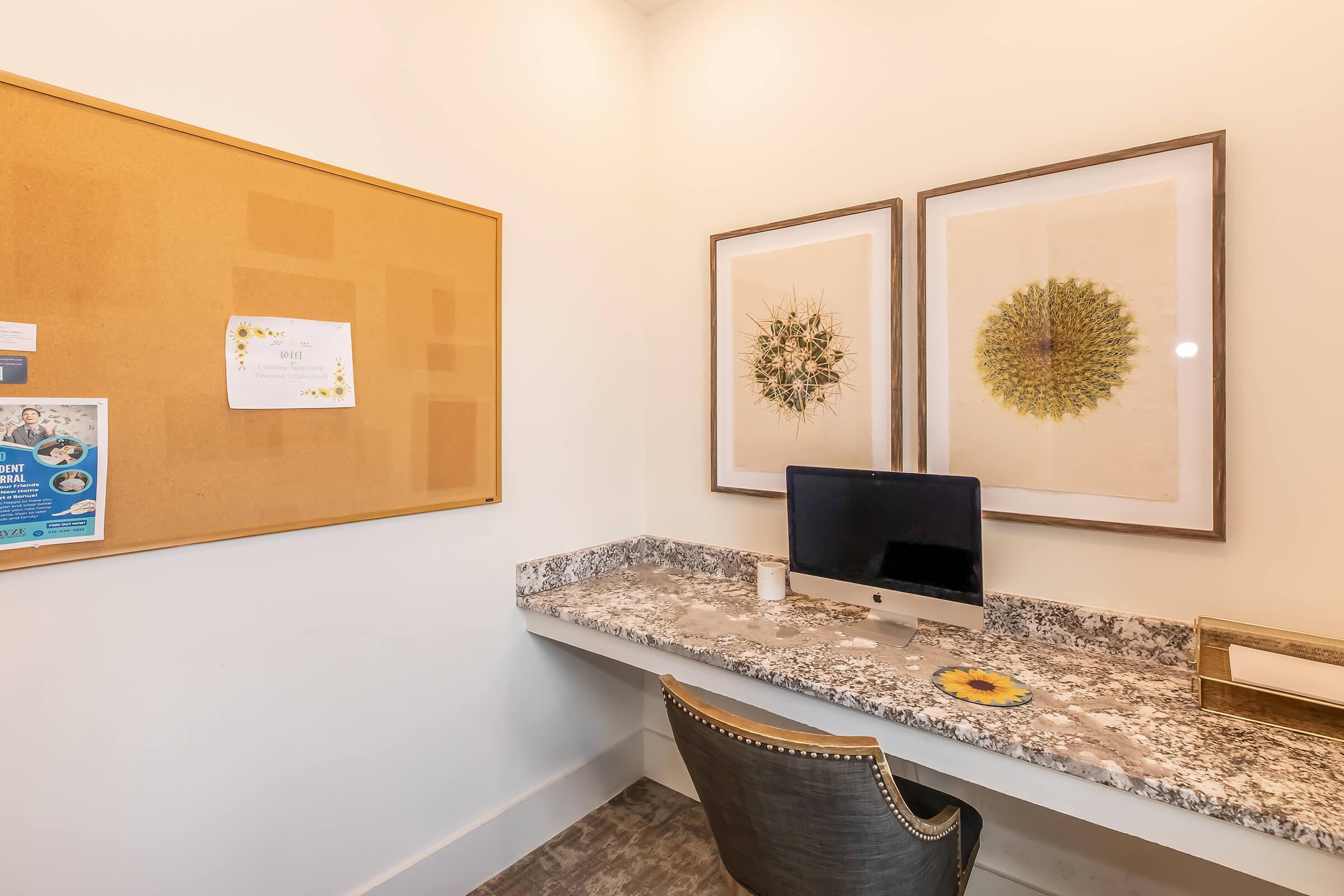 A cozy workspace featuring a granite countertop desk with an iMac computer. The wall displays a bulletin board with notes and a couple of framed botanical prints. A stylish chair is placed at the desk, and a small decorative item sits nearby. The overall ambiance is bright and inviting.