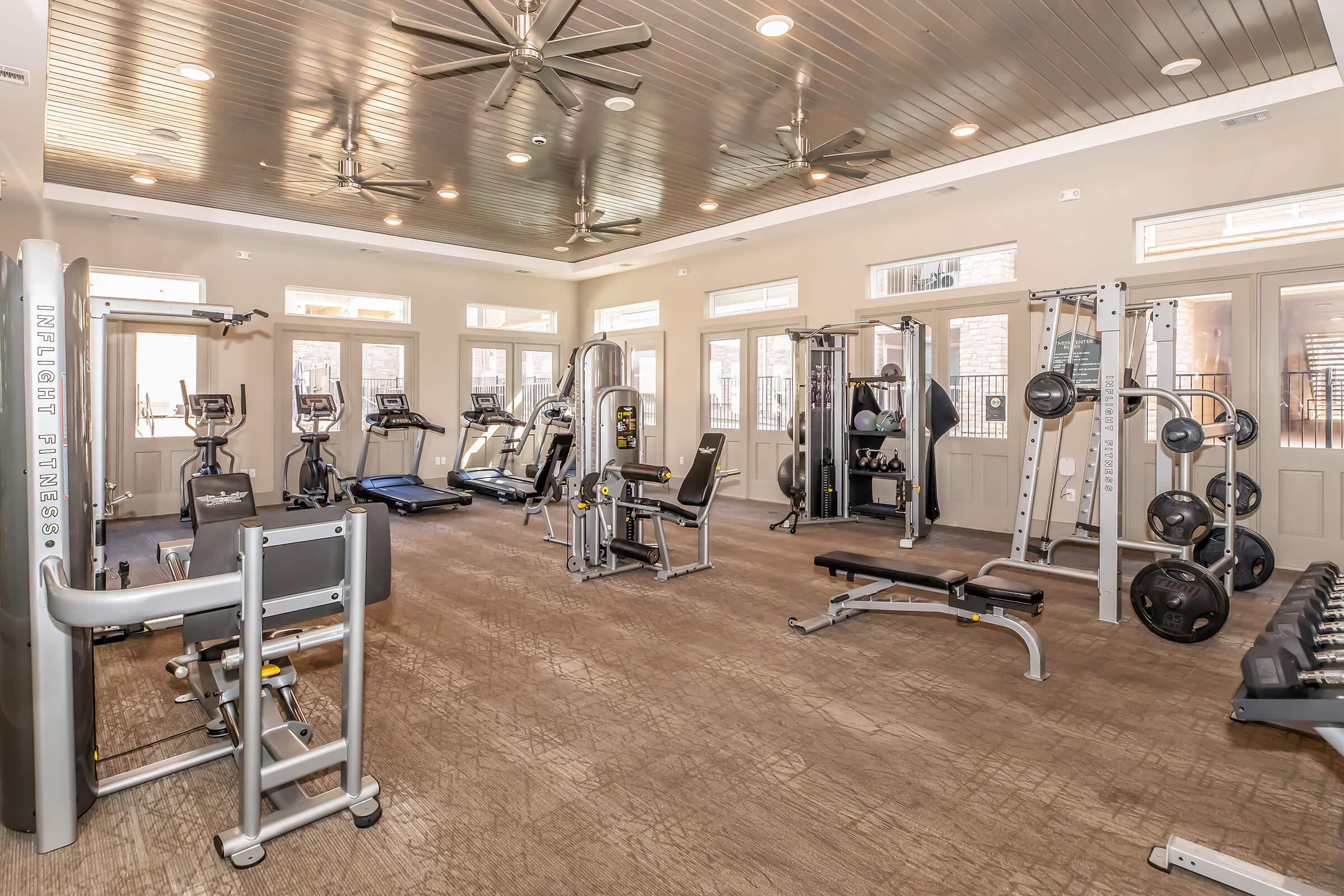 A spacious fitness center featuring various gym equipment, including treadmills, weight machines, free weights, and benches. Large windows allow natural light to fill the room, and ceiling fans provide ventilation, creating an inviting workout environment.