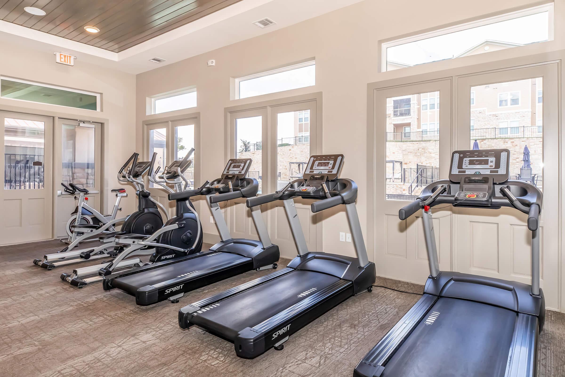 A fitness room with several exercise machines, including treadmills and elliptical trainers, arranged along a wall of large windows. The space is well-lit and designed for workouts, with a clean, modern appearance.