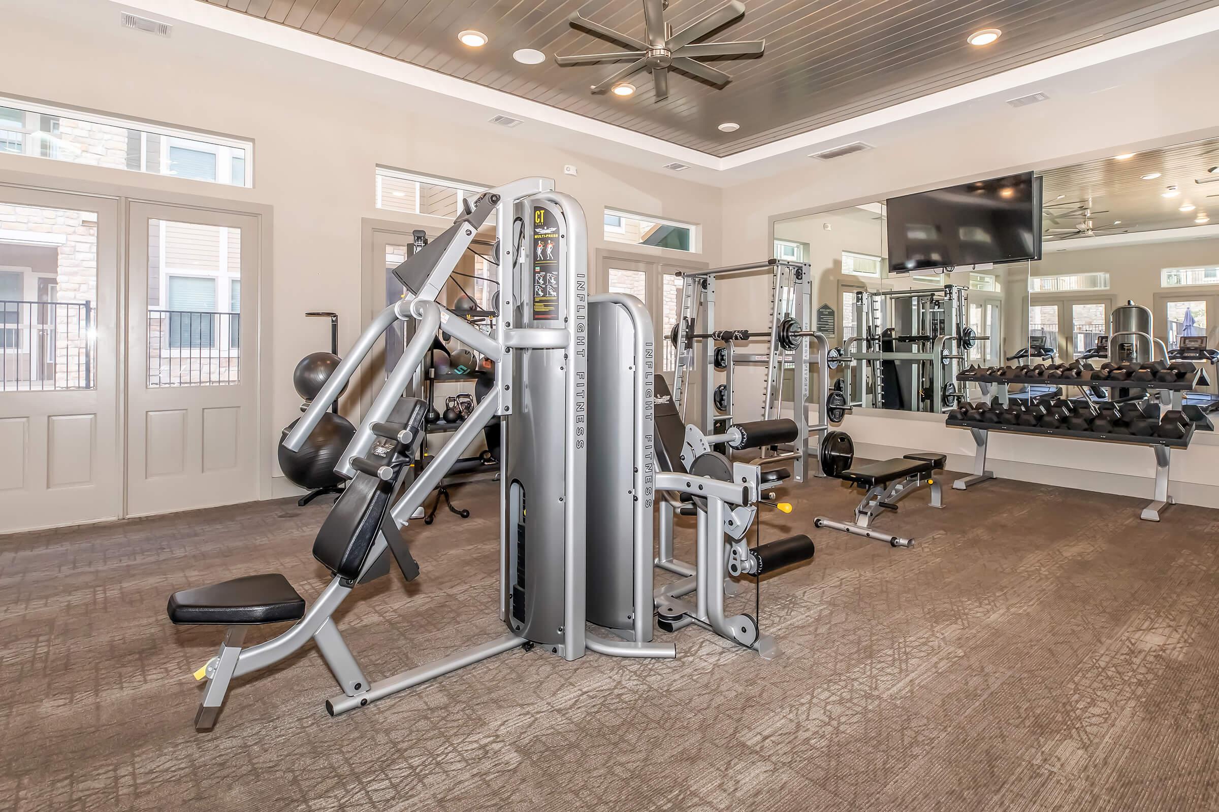 A modern gym interior featuring exercise equipment, including a multi-station machine, free weights, and a large mirror. The space has natural light from windows, a ceiling fan, and is designed for fitness enthusiasts. The floor is carpeted with a neutral tone, creating a welcoming environment.