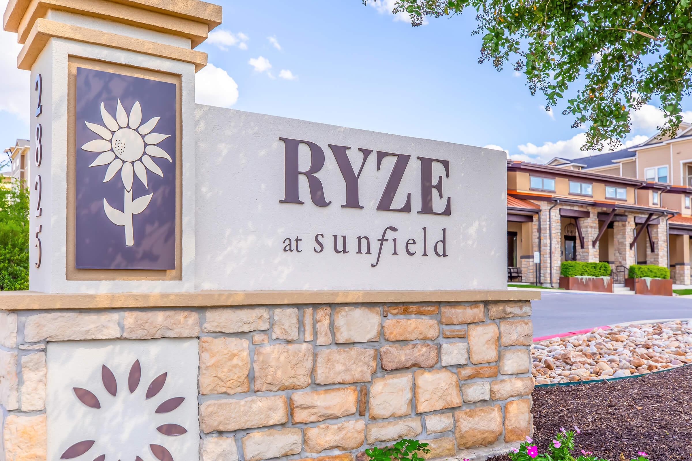 Sign featuring the text "RYZE at Sunfield" with a sunflower graphic. The sign is designed with a combination of stone and decorative elements, surrounded by landscaping and decorative rocks. The setting appears to be a residential or community area, showcasing a modern aesthetic.