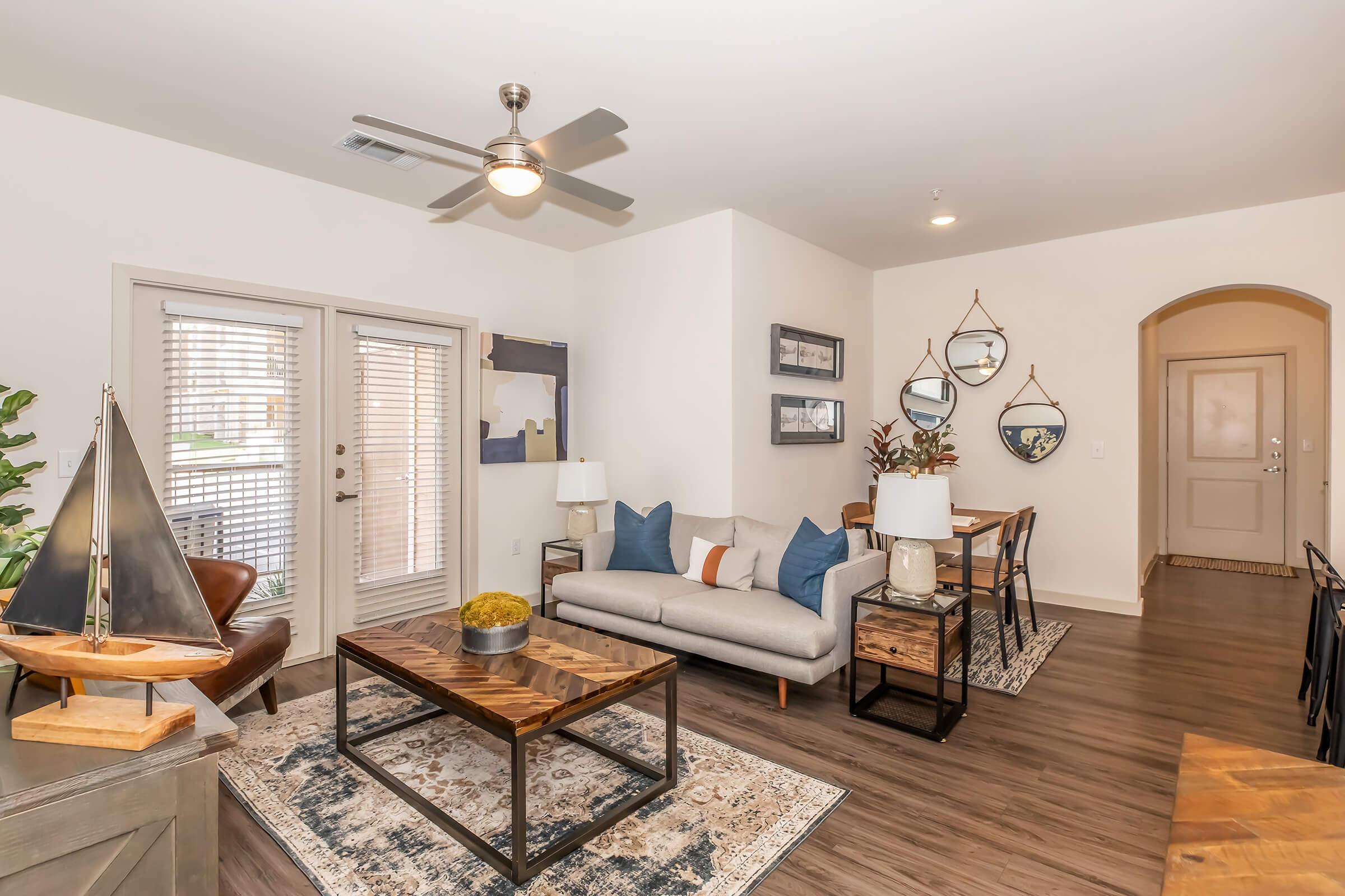 A modern living room featuring a light gray sofa with blue and orange accent pillows, a wooden coffee table, and decorative items. There's a dining area with a wooden table and chairs. Large windows with blinds allow natural light, and there are decorative mirrors and plants enhancing the cozy atmosphere.