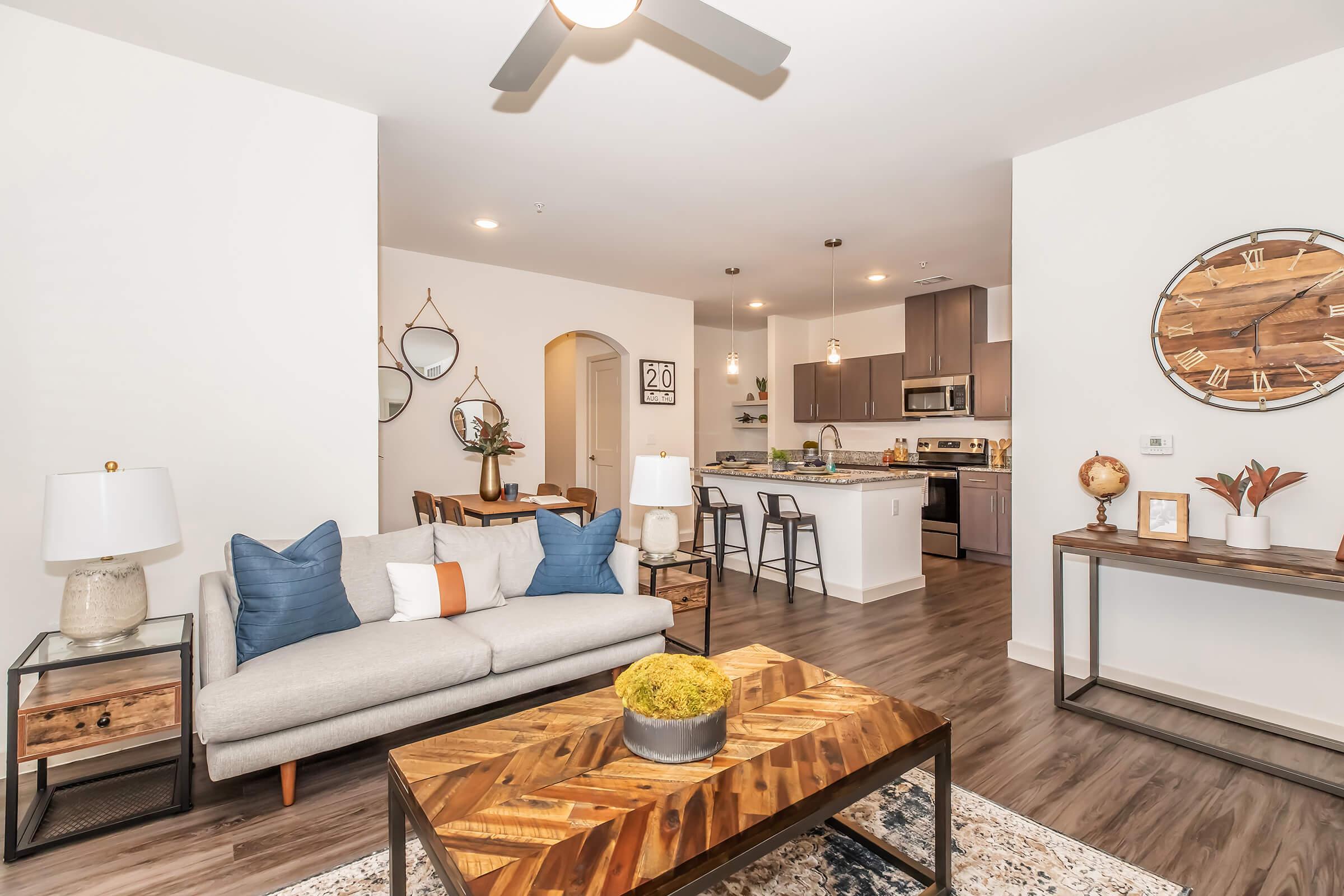 A modern living room featuring a light gray sofa with blue and orange pillows, a wooden coffee table with decorative items on top, and a stylish area rug. There are decorative mirrors on the wall, a dining area in the background with bar stools, and a contemporary clock on the wall, creating a warm and inviting atmosphere.