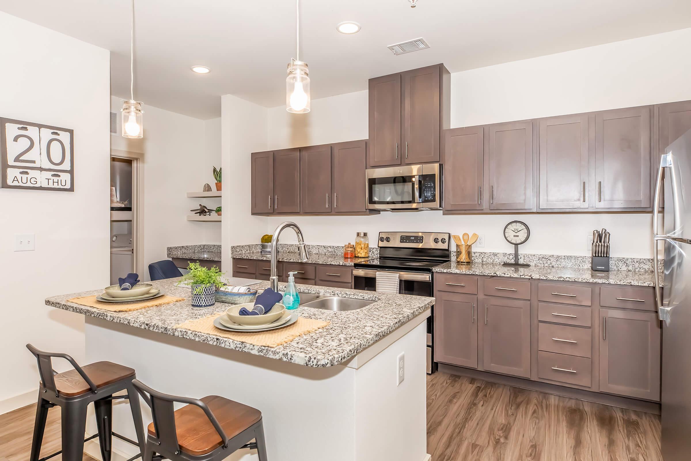 Modern kitchen featuring dark wooden cabinets and granite countertops. The island has two bar stools with decorative table settings. Stainless steel appliances, including a microwave and oven, are visible. Pendant lighting hangs above the island, and a clock is mounted on the wall. Bright and inviting atmosphere.