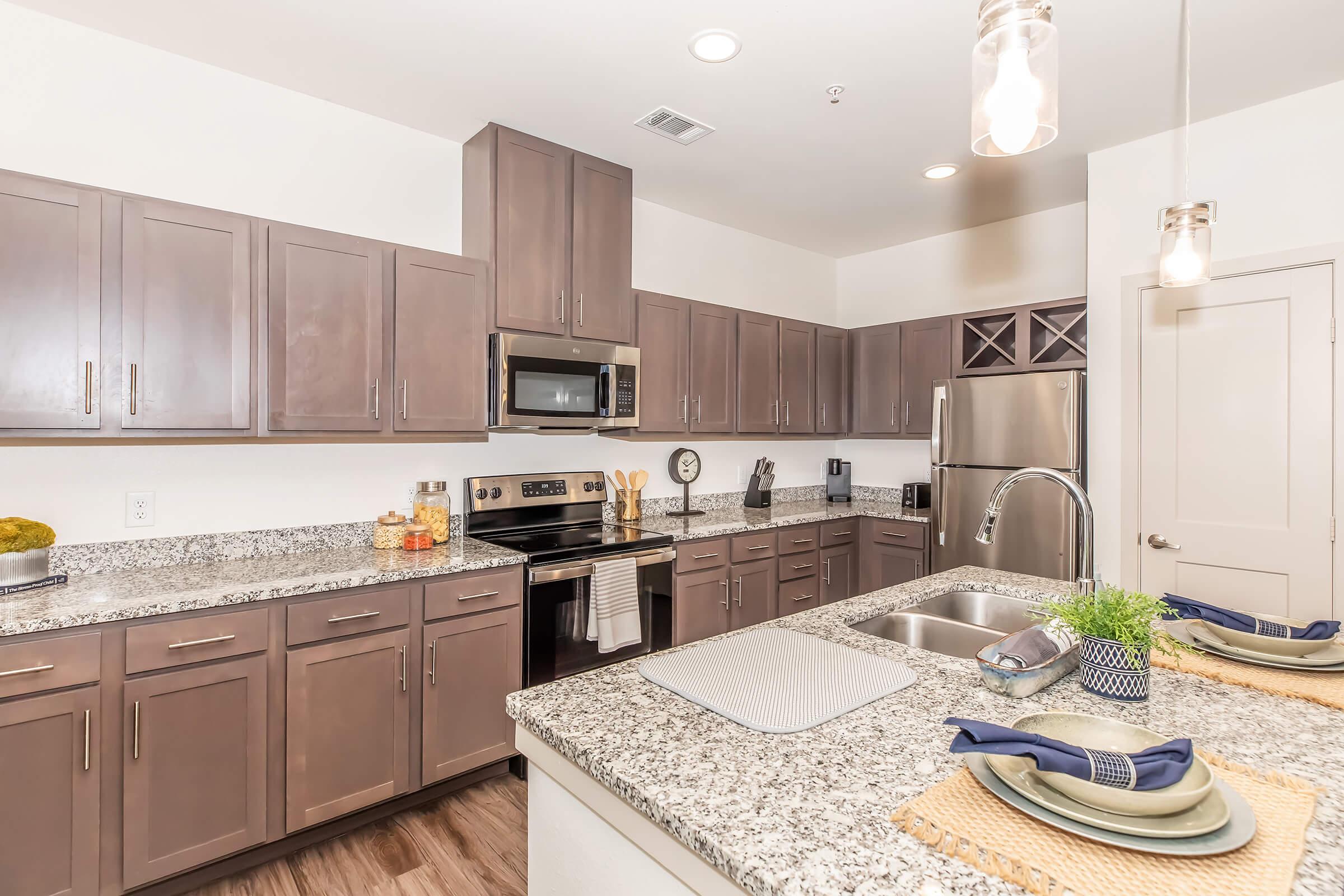 Modern kitchen with dark wood cabinets and granite countertops. Stainless steel appliances, including a microwave and dishwasher, are visible. A sink with a modern faucet is situated on an island with seating. Decorative items like fruits and a small plant add a touch of style. Soft lighting illuminates the space.