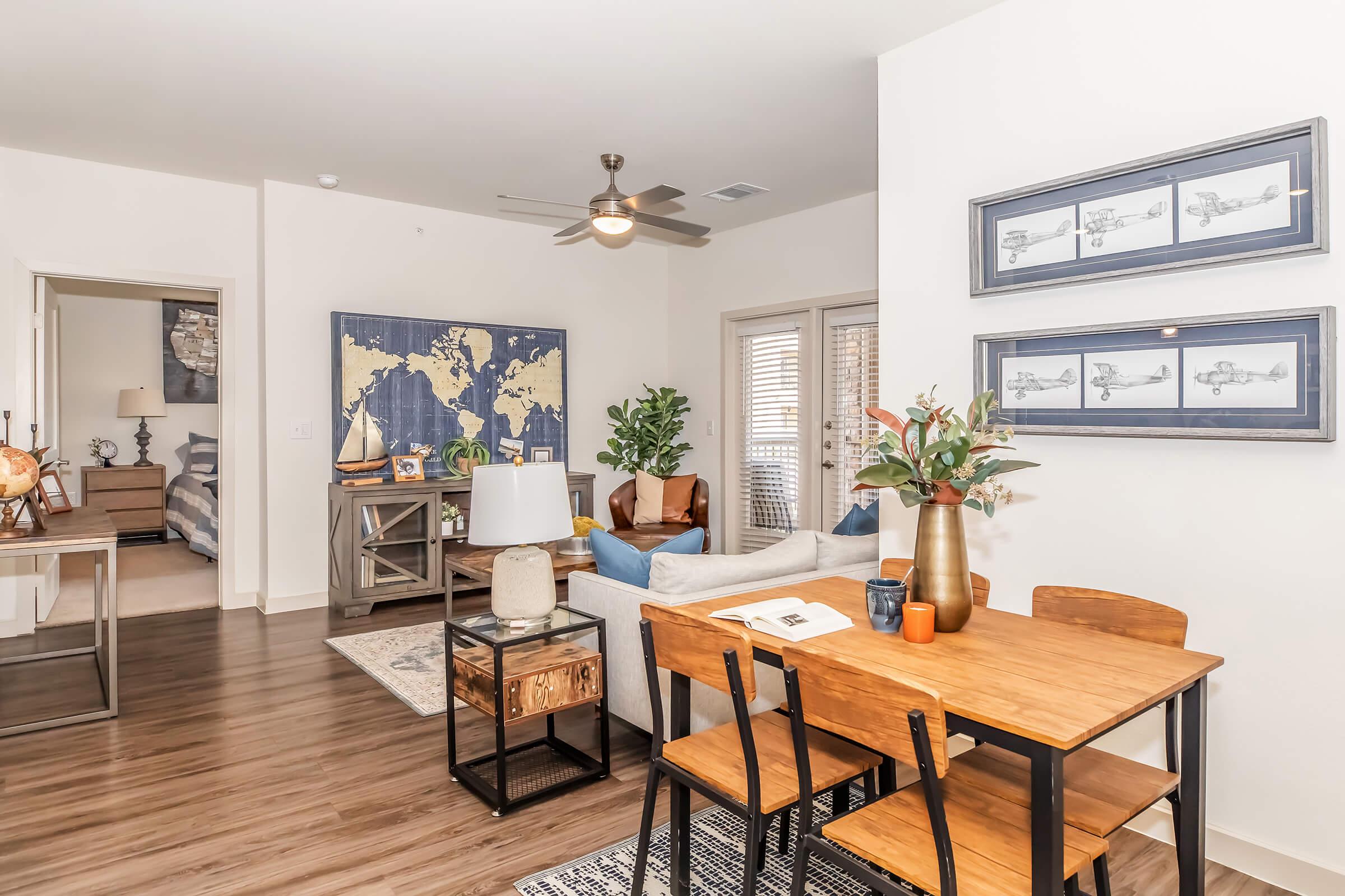 A well-decorated living space featuring a dining area with a wooden table and chairs, a cozy sitting area with a blue sofa, and a world map on the wall. There are plants, decorative items, and a ceiling fan, creating a warm and inviting atmosphere in a contemporary apartment.