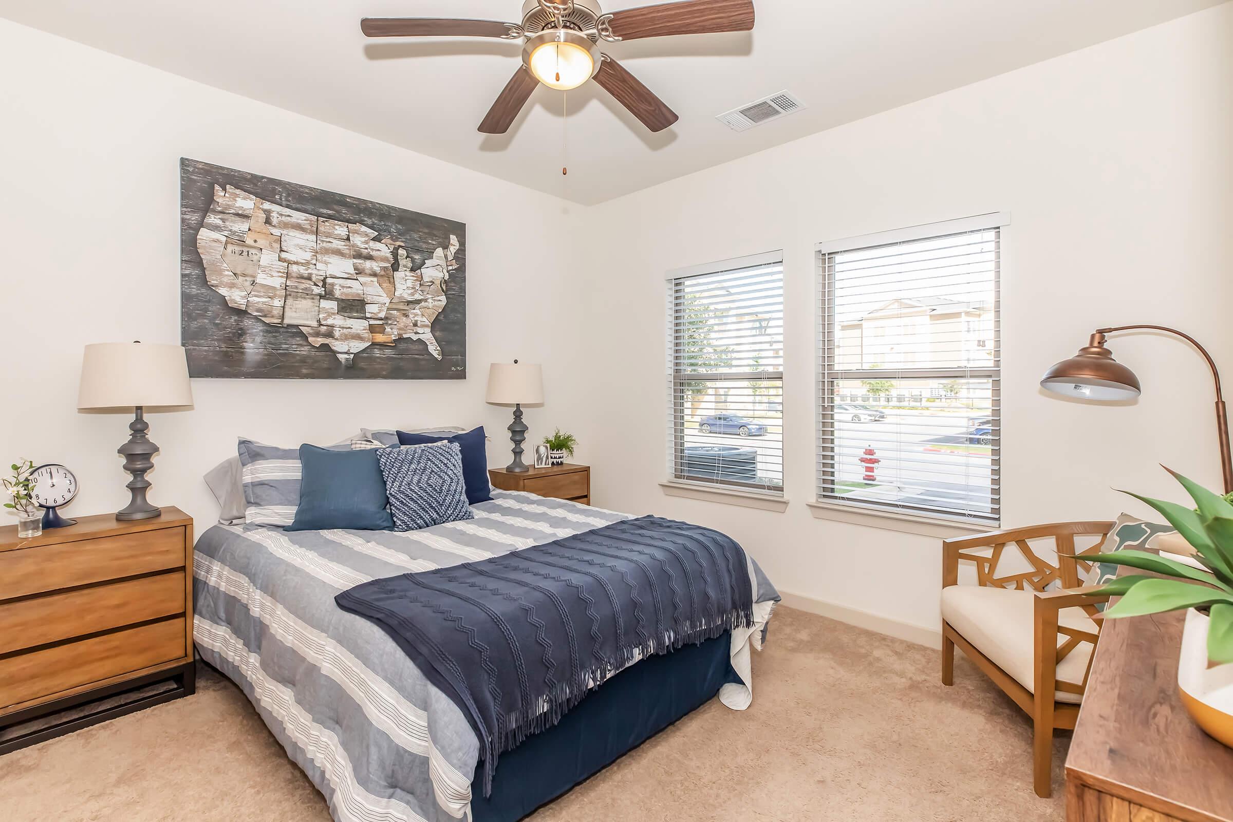 A cozy bedroom featuring a bed with blue and gray striped bedding, two bedside lamps on wooden nightstands, a large wall art piece of a United States map, a ceiling fan, a comfortable chair, and two windows with blinds allowing natural light.