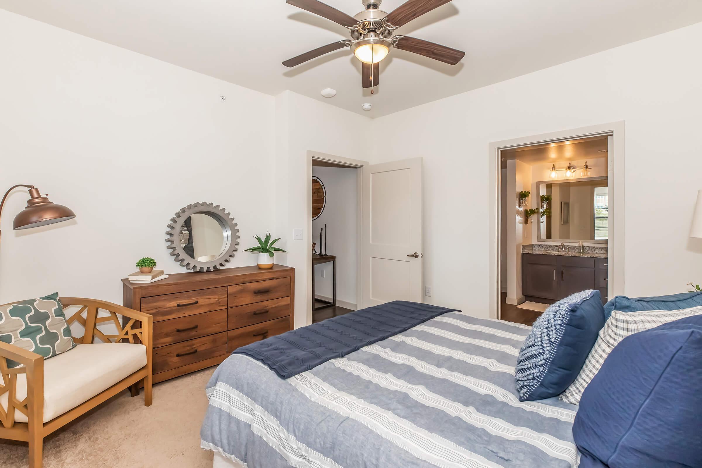 A cozy bedroom featuring a striped blue and white bedspread, a wooden dresser, a decorative mirror, and a chair with patterned cushions. A ceiling fan provides airflow, and there is a view of an adjoining bathroom with a double sink in the background. Natural light enhances the warm atmosphere.