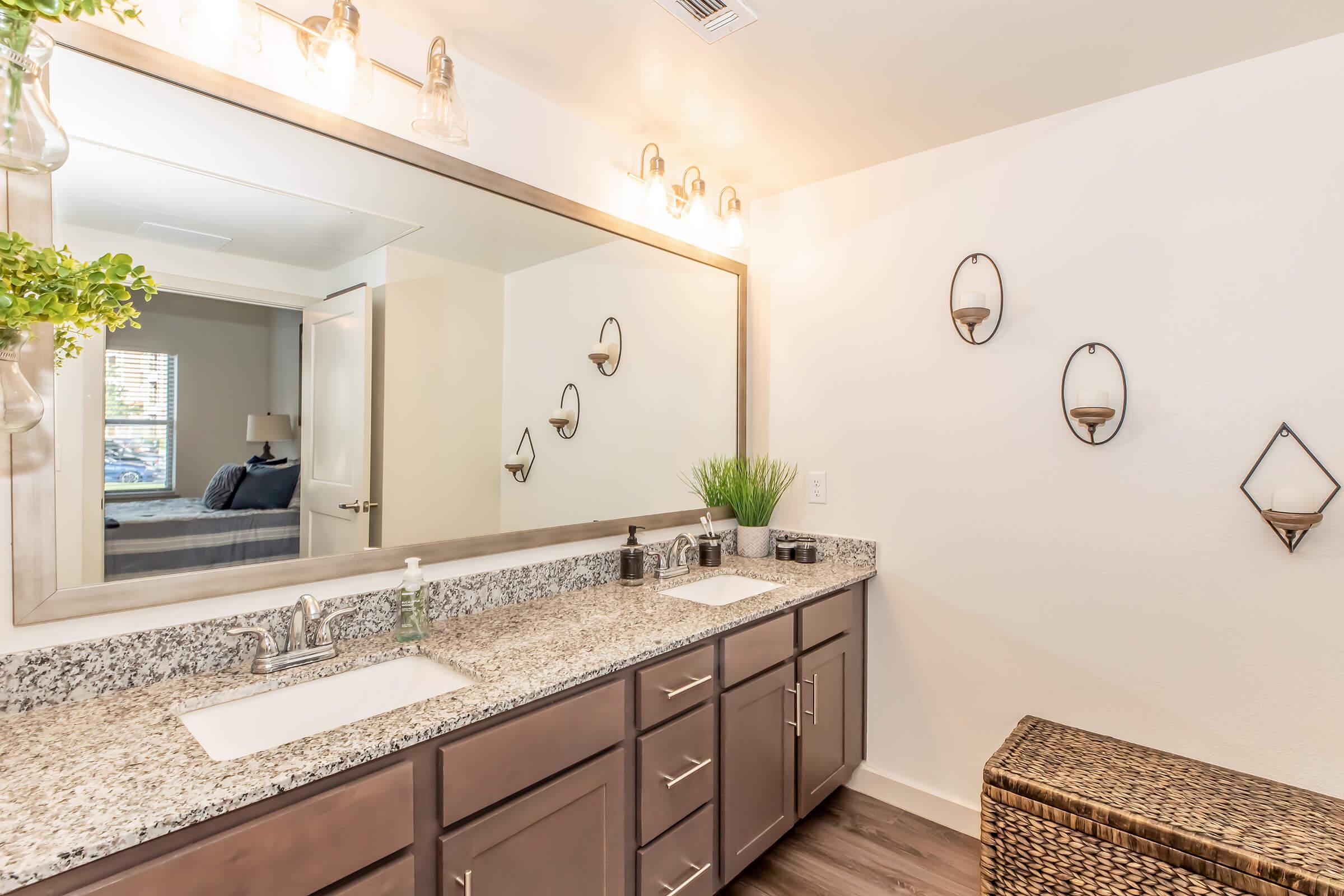 A modern bathroom featuring a spacious countertop with two sinks, a large mirror with stylish lighting above, and decorative wall sconces. A woven basket sits beside the sinks, and a touch of greenery adds a fresh element. The background indicates a cozy bedroom area.