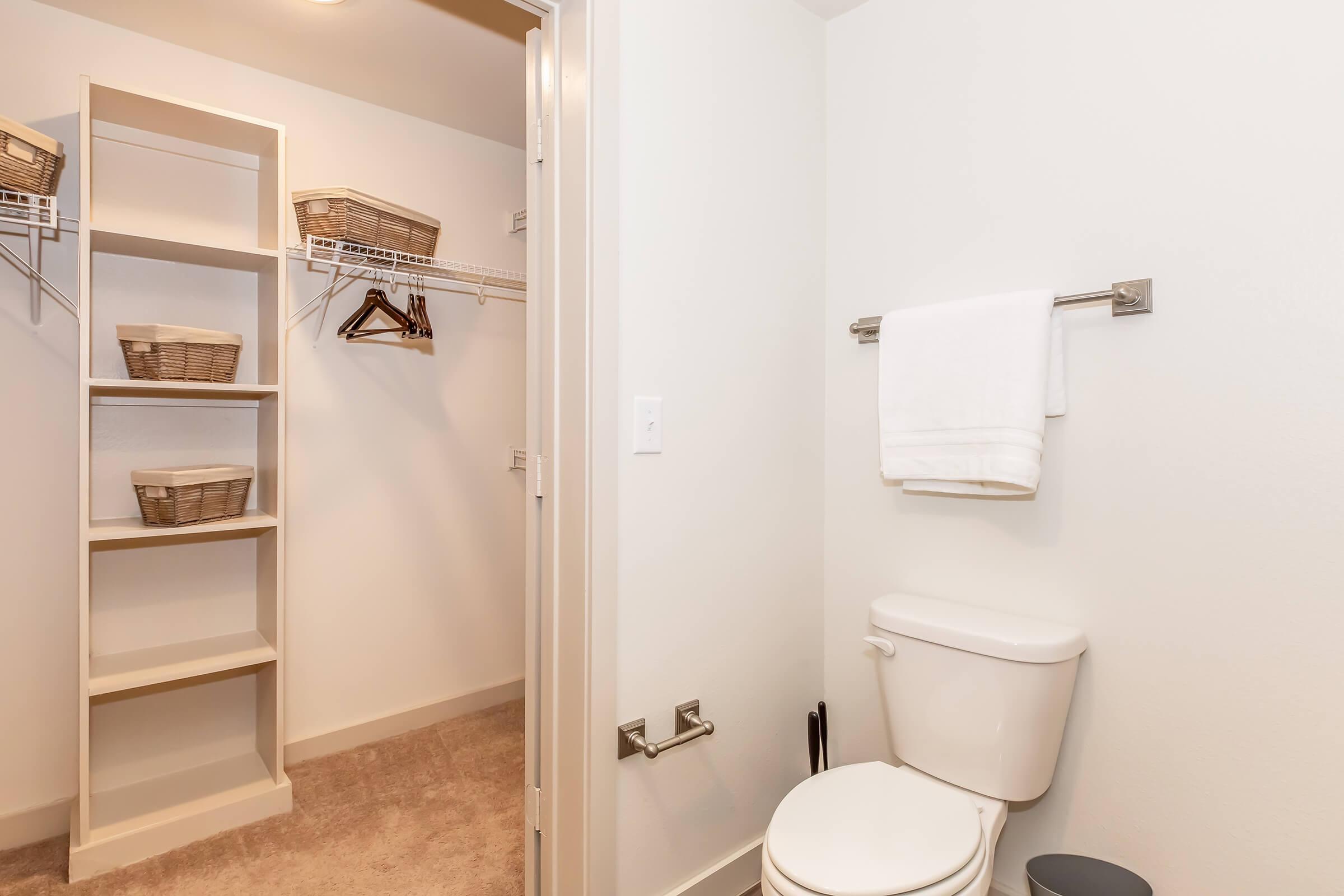 A clean bathroom featuring a toilet next to a small closet with open shelves. The closet has wicker baskets and hangers for storage. A white towel is hung on a towel rack, and the walls are painted in a light color, creating a bright and simple aesthetic. The floor is carpeted for added comfort.