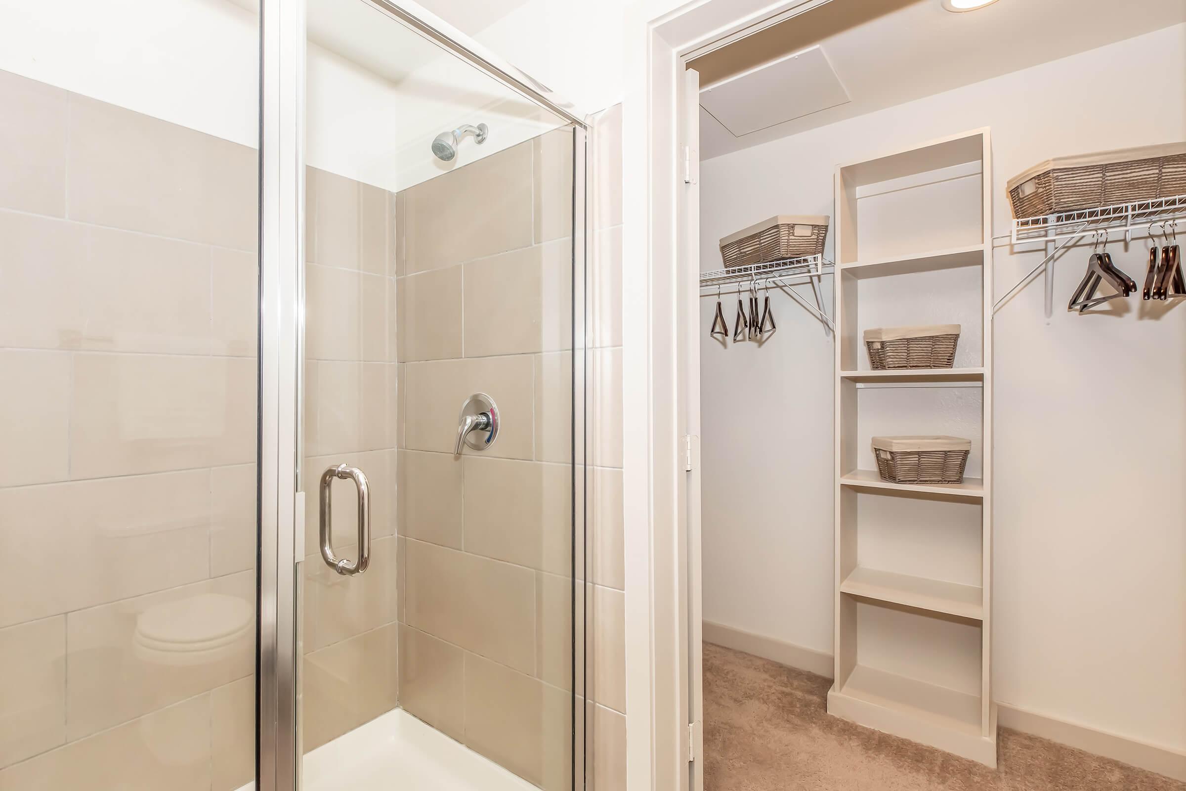 A modern bathroom featuring a glass shower stall with tiled walls. Adjacent is a small walk-in closet with shelves and baskets for storage, equipped with hangers. The space is bright and uncluttered, showcasing a neutral color palette with soft lighting.