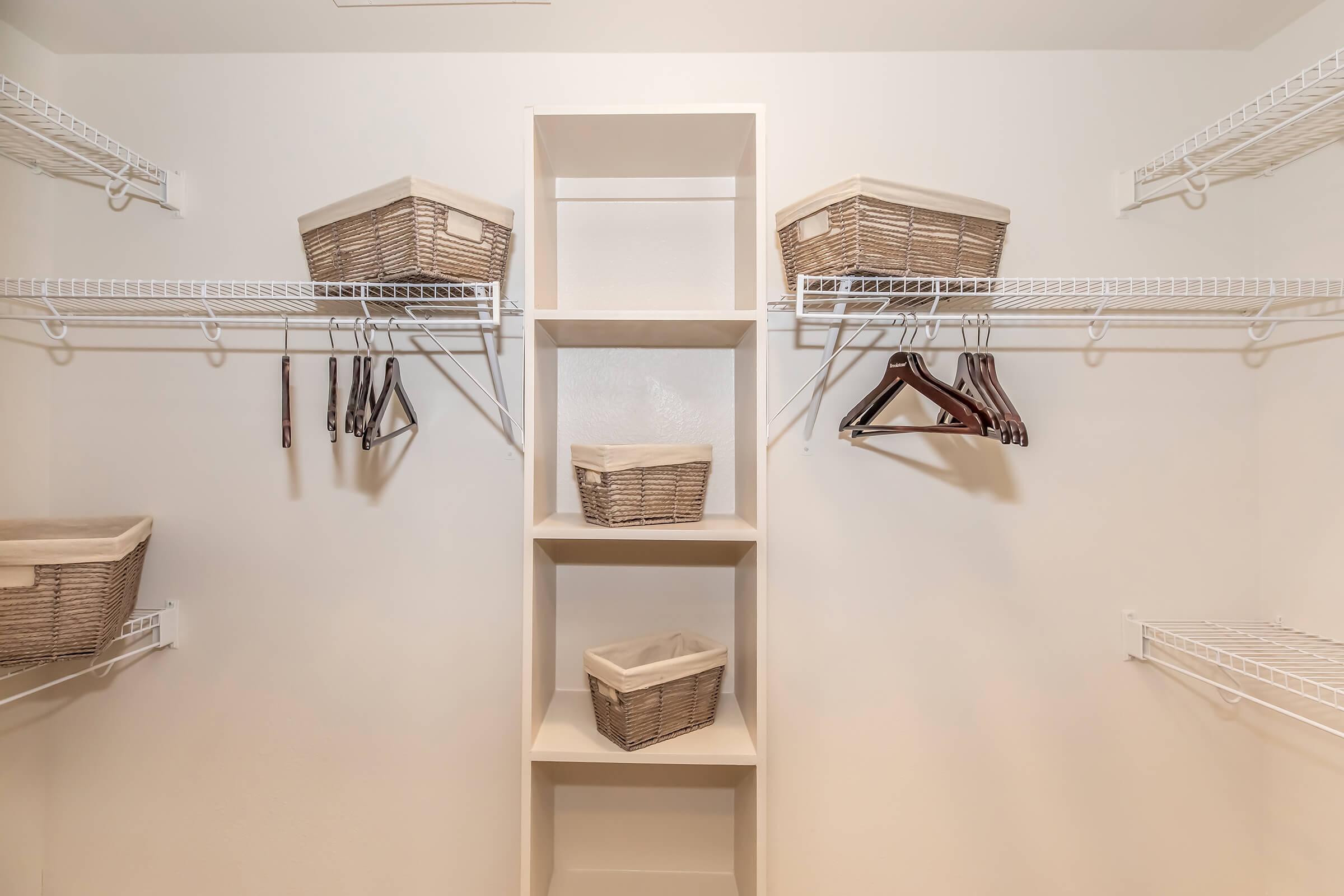 A tidy closet interior featuring white wire shelves. There are three woven storage baskets on the upper shelves and two hooks with hangers on the sides. A central open shelf provides additional storage space, all set against a plain, light-colored wall.