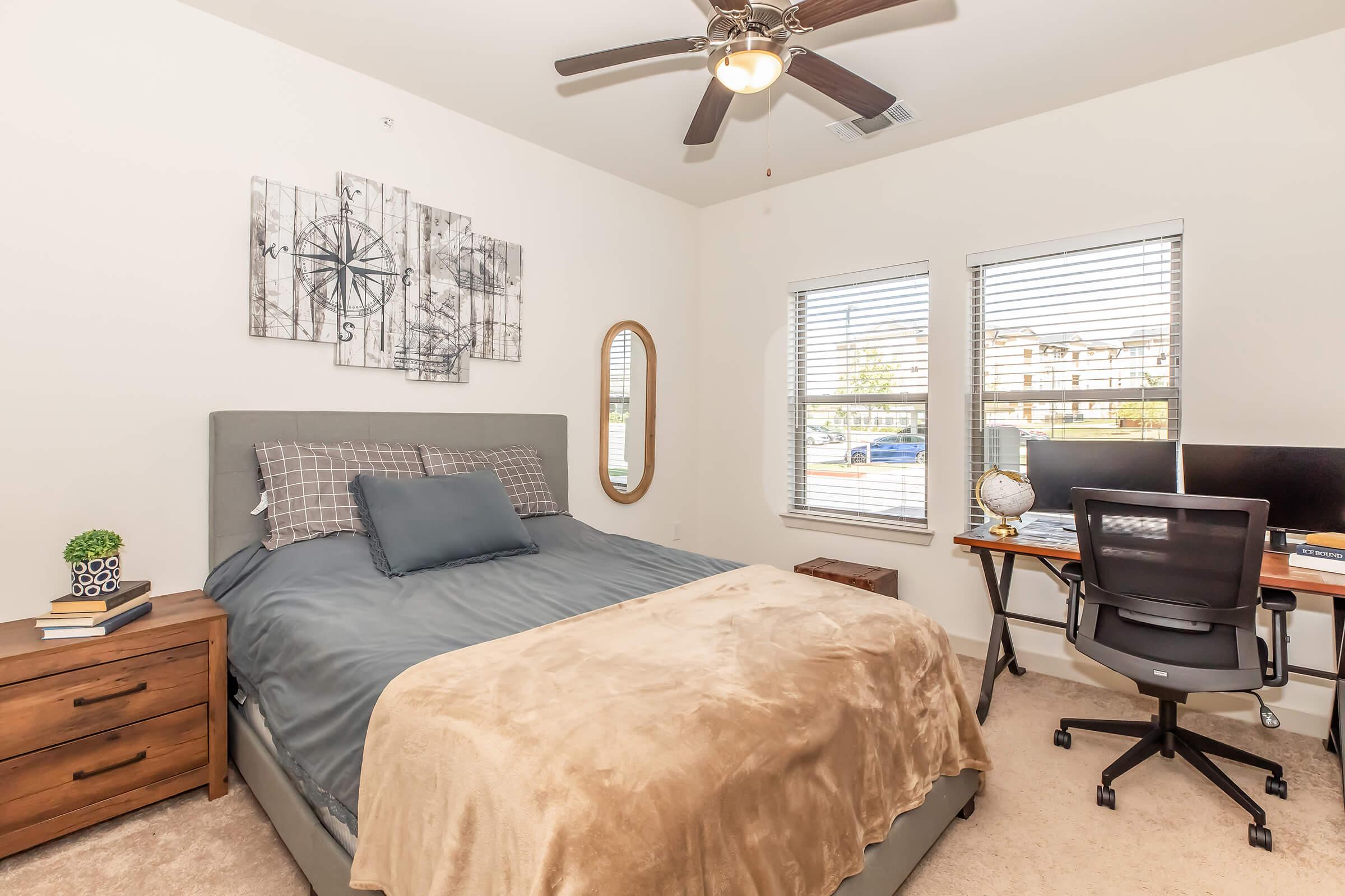 A cozy bedroom featuring a gray upholstered bed with a blue blanket, a wooden nightstand, and a round mirror on the wall. There are two windows with blinds, offering natural light, and a desk with two monitors set up in the corner, creating a functional workspace.