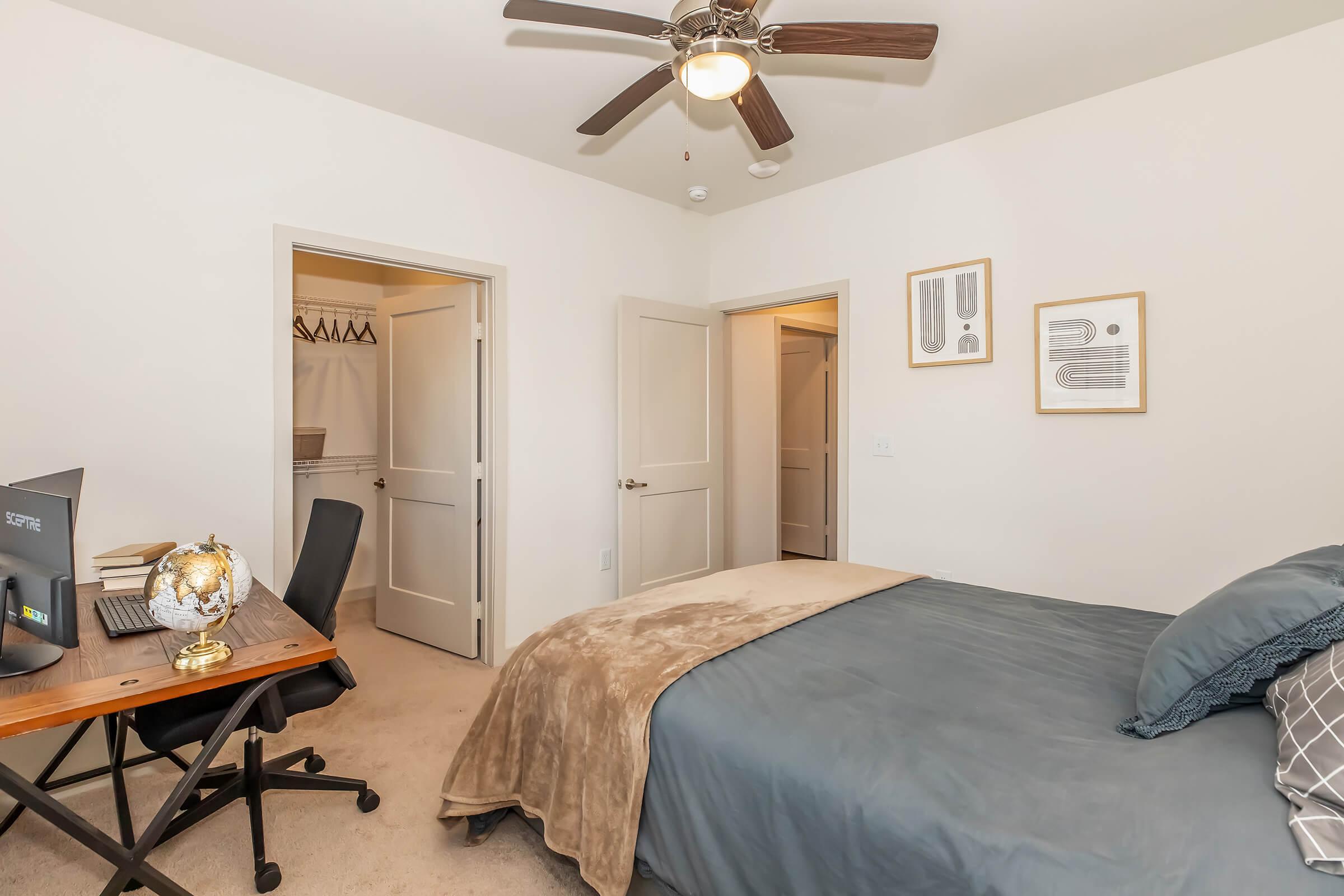 A cozy bedroom featuring a neatly made bed with gray bedding, a wooden desk with a globe and a black chair, and a ceiling fan. Two doors leading to a closet and another room are visible, along with framed wall art. The room has neutral-colored walls and carpeted flooring.