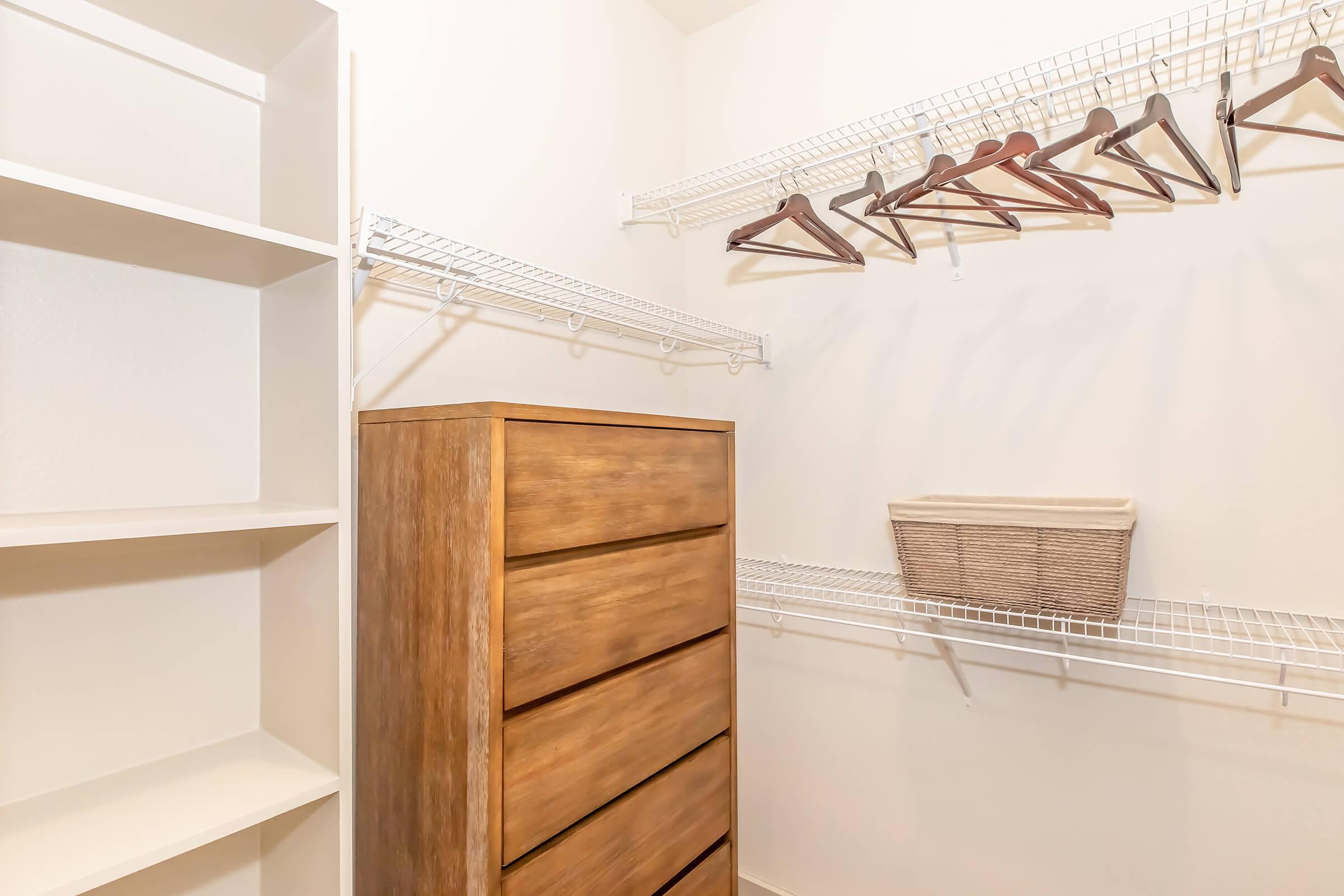 A neatly organized closet featuring a wooden dresser with five drawers, open shelves on the left, and wire shelving above with several hangers. A woven basket sits on the wire shelf, all set against a plain white wall for a clean, minimalist look.