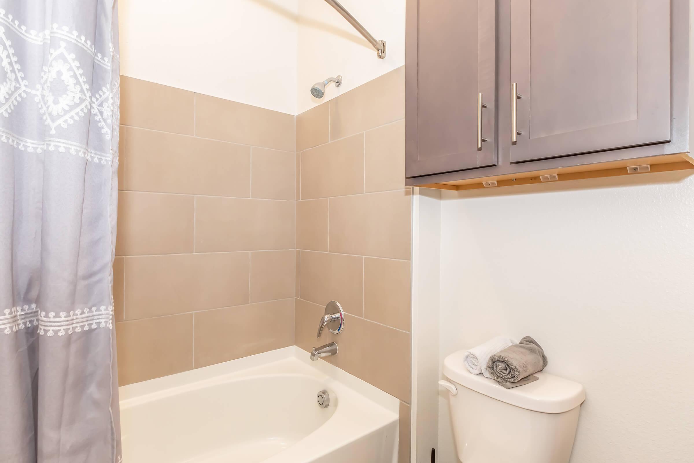 A modern bathroom featuring a bathtub with a shower curtain, light-colored tile walls, a chrome showerhead, and a wall-mounted cabinet above a toilet. Neatly rolled towels are placed on a shelf next to the toilet. The overall design is clean and minimalist.