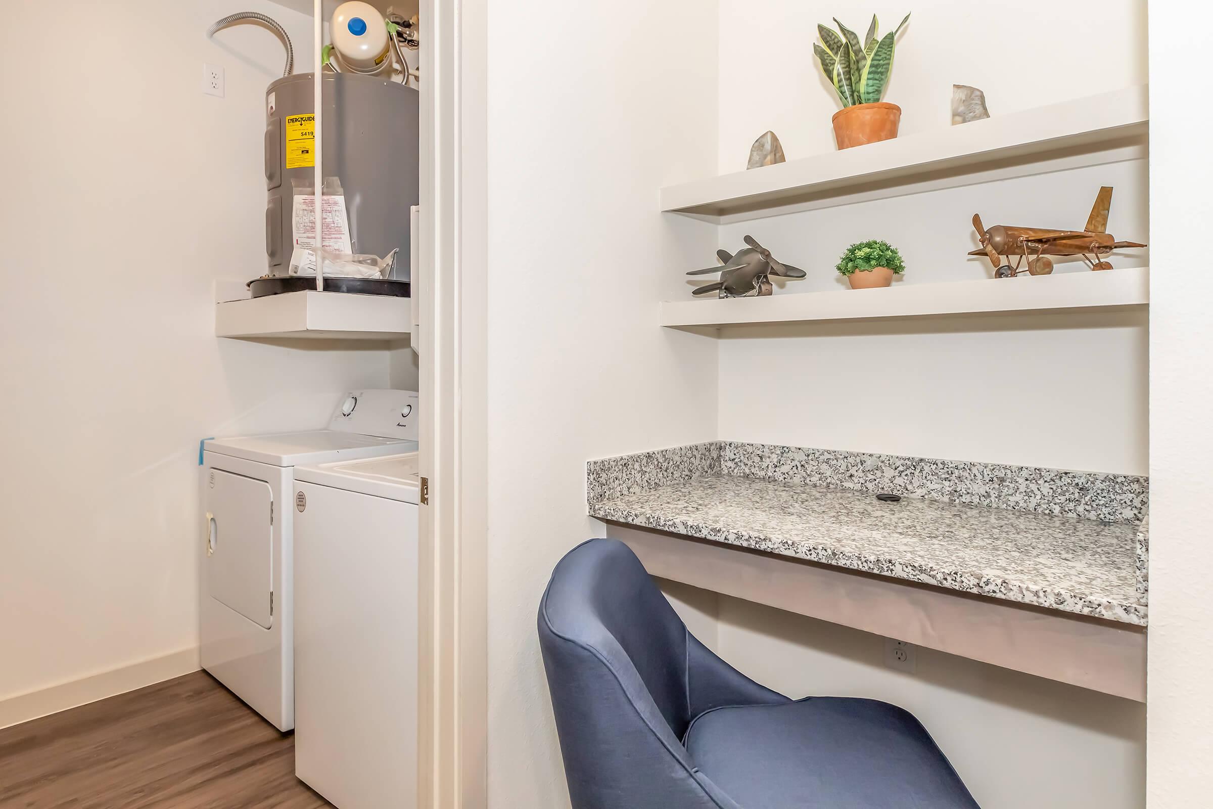 A small laundry area featuring a stacked washer and dryer next to a gray wall. A granite countertop workspace with a blue chair is in the foreground. Above the counter, there are shelves displaying decorative items like small plants and model airplanes. The space is well-lit and modern.