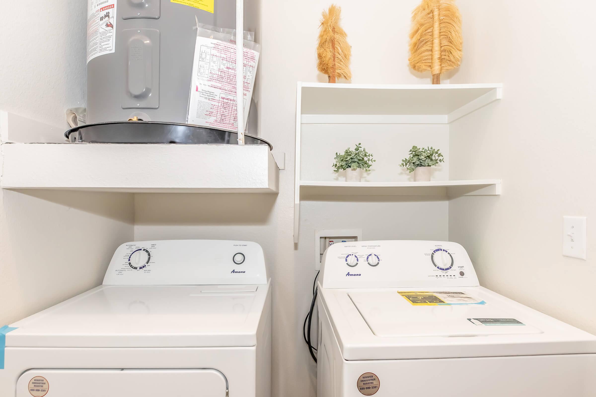 A clean laundry room featuring a stacked washer and dryer in white. Above the appliances, there is a water heater and a shelf with decorative items, including two small green plants and a pair of yellow feather-like decorations. The walls are painted in a light color, creating a bright atmosphere.
