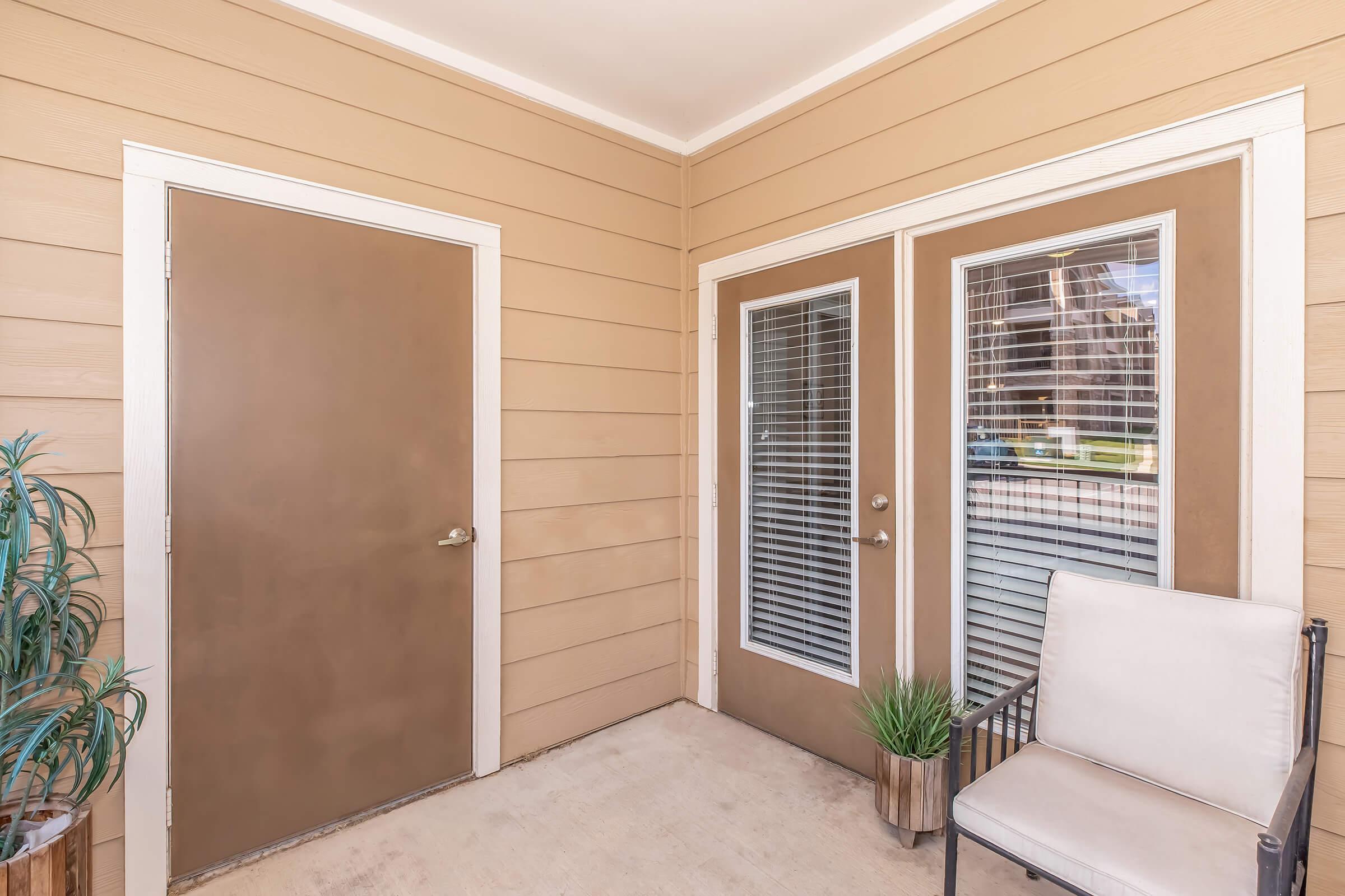 An entryway featuring a closed brown door and two glass-paneled doors with blinds, leading to an outdoor area. There's a small potted plant beside the doors and a single metal chair positioned on a beige surface. The walls are a light tan color, creating a warm and inviting atmosphere.