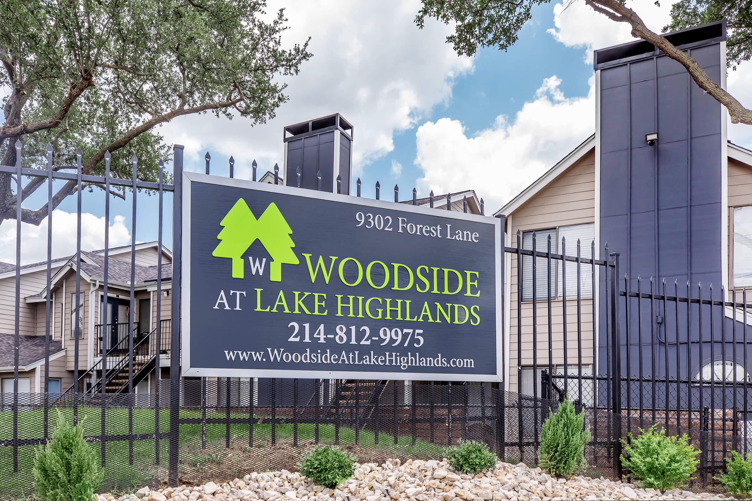 Sign for "Woodside at Lake Highlands" located at 9302 Forest Lane, featuring a black and green design with the property name, phone number, and website. The sign is surrounded by a fence, with landscaped stone and small plants in the foreground, and a building visible in the background.