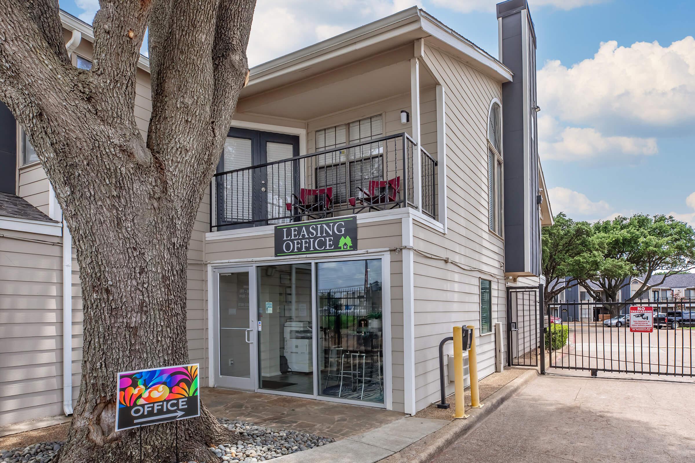 Exterior view of a leasing office building, featuring a sign that reads "Leasing Office." The office has a balcony with two chairs and is surrounded by trees. There is a colorful sign near the entrance, and a gate leading to a parking area is visible in the background. The sky is blue with scattered clouds.