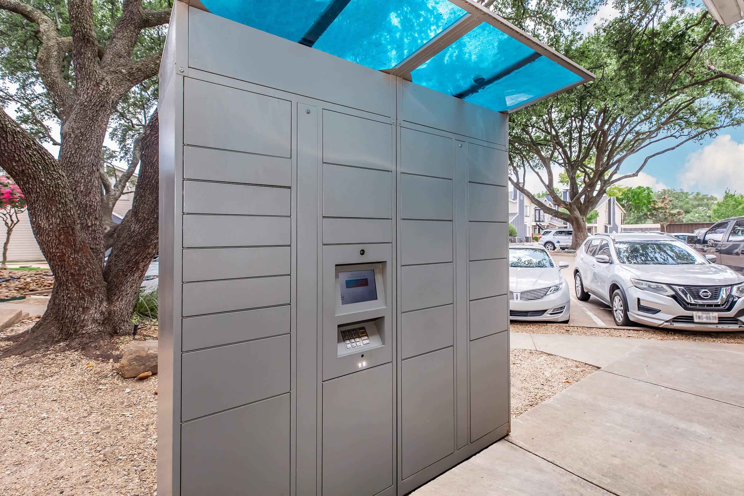 A gray package delivery locker kiosk located in an outdoor area, surrounded by trees and parked cars. The structure features a transparent blue roof and multiple compartments for retrieving parcels. The setting is well-lit with a clear sky in the background.