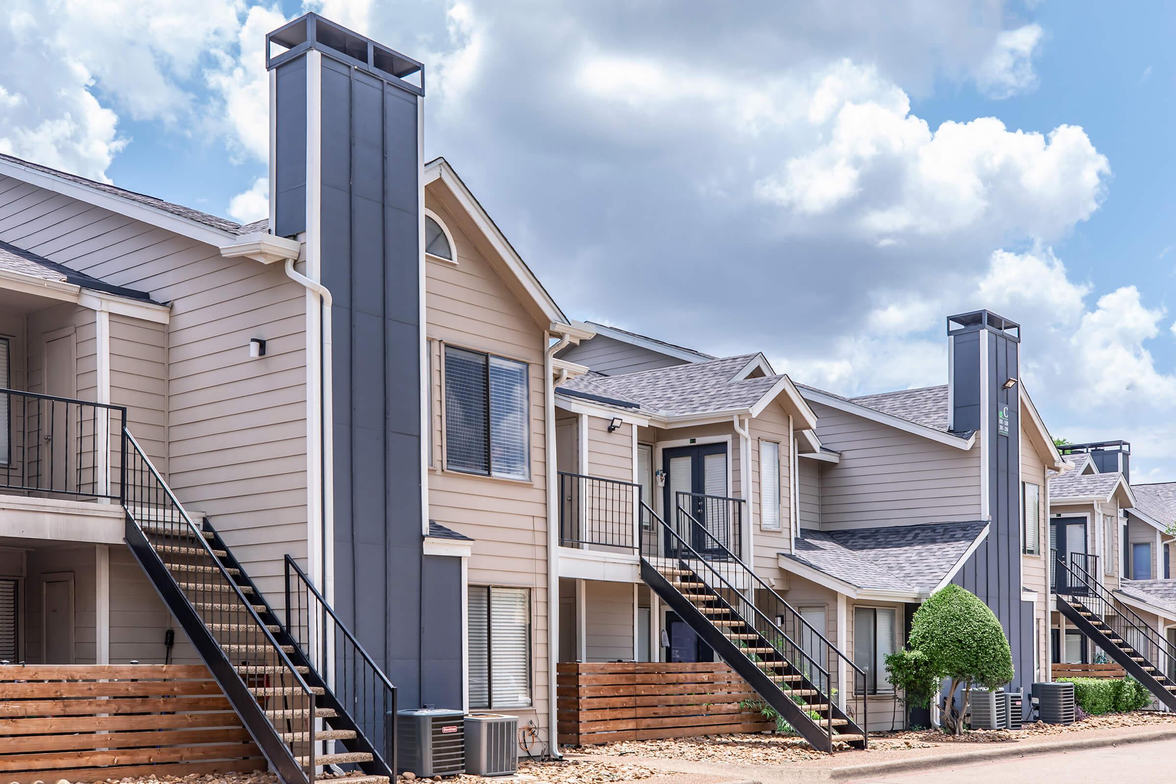 A row of modern apartment buildings featuring two-story structures with balconies, gray and beige siding, and prominent vertical chimneys. The buildings are surrounded by landscaped areas and a clear blue sky with fluffy white clouds.