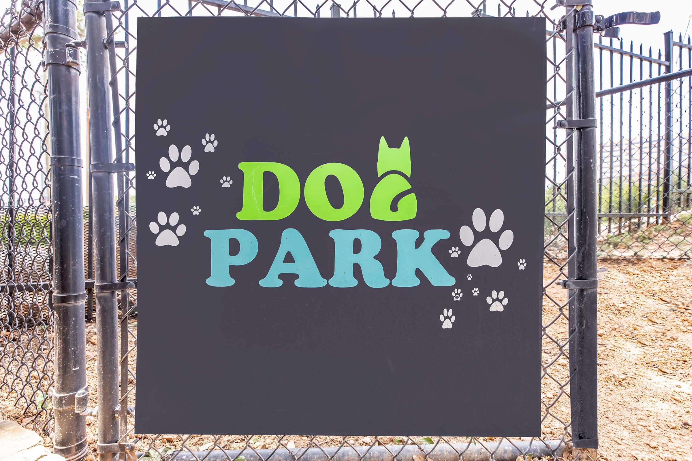 Sign for a dog park featuring the words "DOG PARK" in large, colorful letters. The word "DOG" is green with a playful font, and "PARK" is in light blue. Surrounding the text are various paw prints, adding to the playful theme. The sign is mounted on a fence in an outdoor setting.