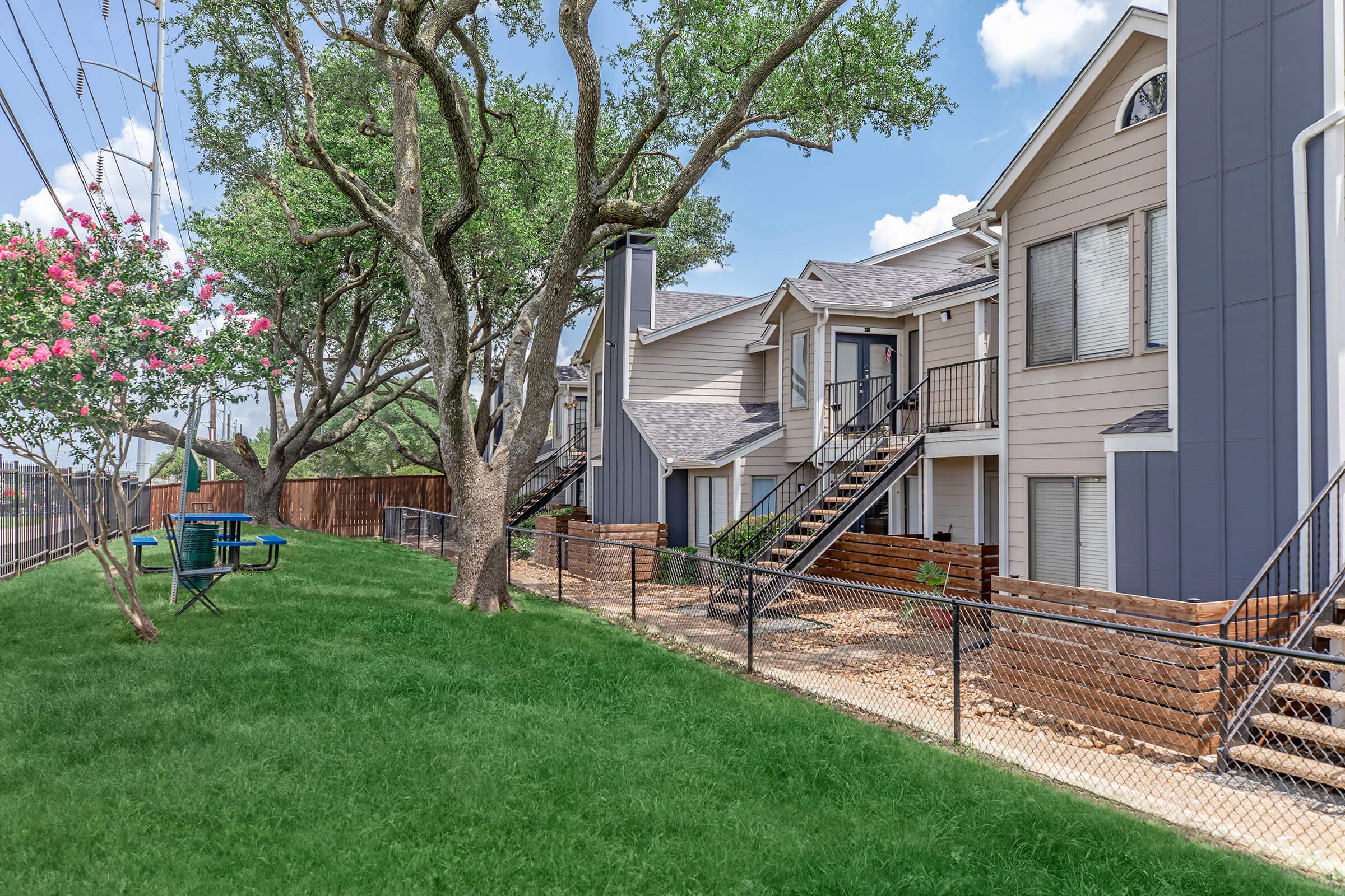 A well-maintained residential area featuring multi-story apartment buildings with balconies, surrounded by green grass and trees. There are benches and picnic tables in the yard, and a fence runs along the property boundary. The sky is clear with a few clouds.