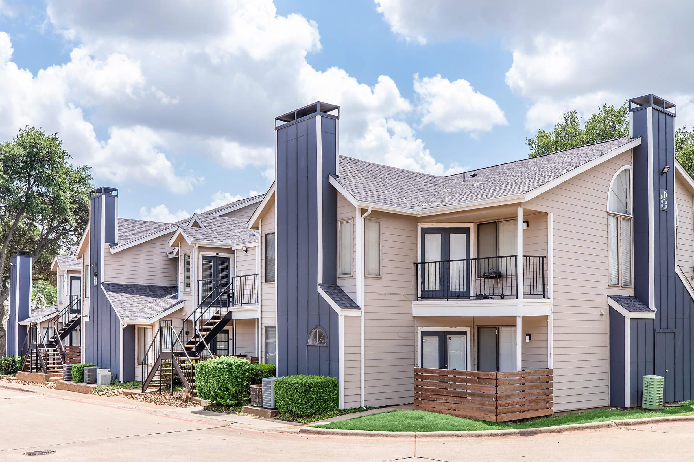 A row of modern multi-unit apartments with a mix of light and dark blue siding. Each unit features a balcony, large windows, and a staircase leading to the upper units. The landscape includes manicured shrubs and a clear blue sky with fluffy clouds.