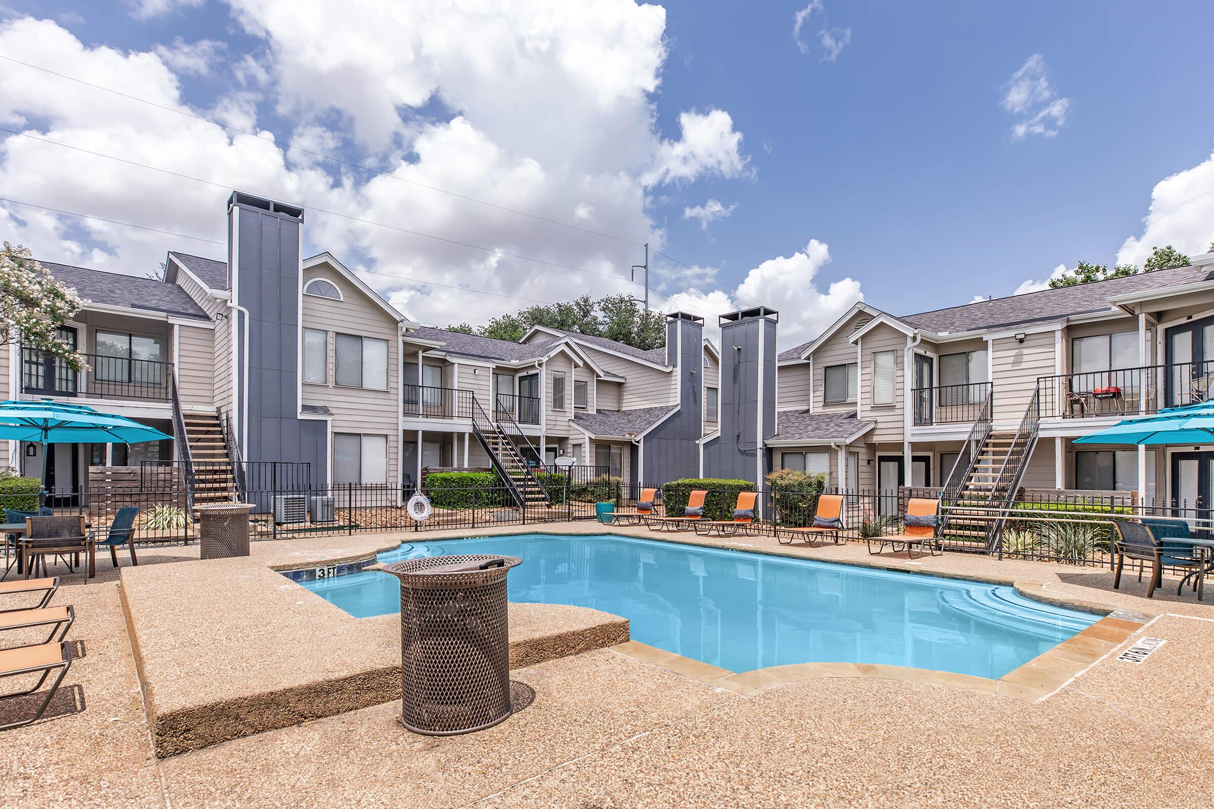 A well-maintained apartment complex courtyard with a clear blue pool at the center. Surrounding the pool are lounge chairs and umbrellas, with two-story apartment buildings in the backdrop. The sky is partly cloudy, creating a bright and inviting atmosphere.