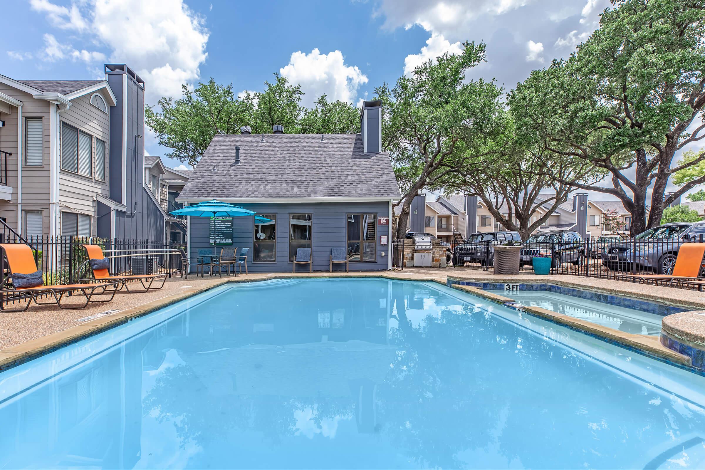 A clear blue swimming pool surrounded by lounge chairs and umbrellas, with a residential building in the background. Lush green trees provide shade, and a few parked cars are visible nearby. The sky is bright with scattered clouds, creating a serene outdoor atmosphere.