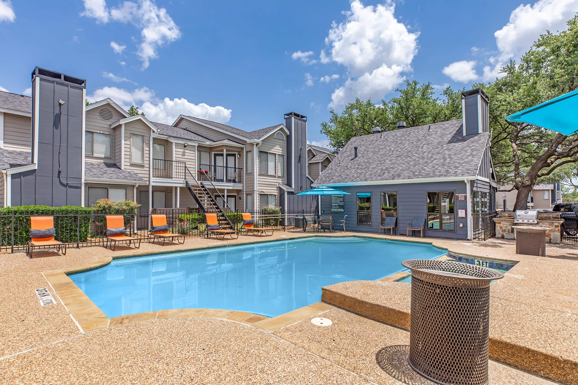 A bright, sunny day at an apartment complex featuring a sparkling blue swimming pool surrounded by lounge chairs. The area includes barbecuing facilities, greenery, and well-maintained buildings in the background, creating a relaxing outdoor atmosphere. Fluffy clouds drift across the blue sky.