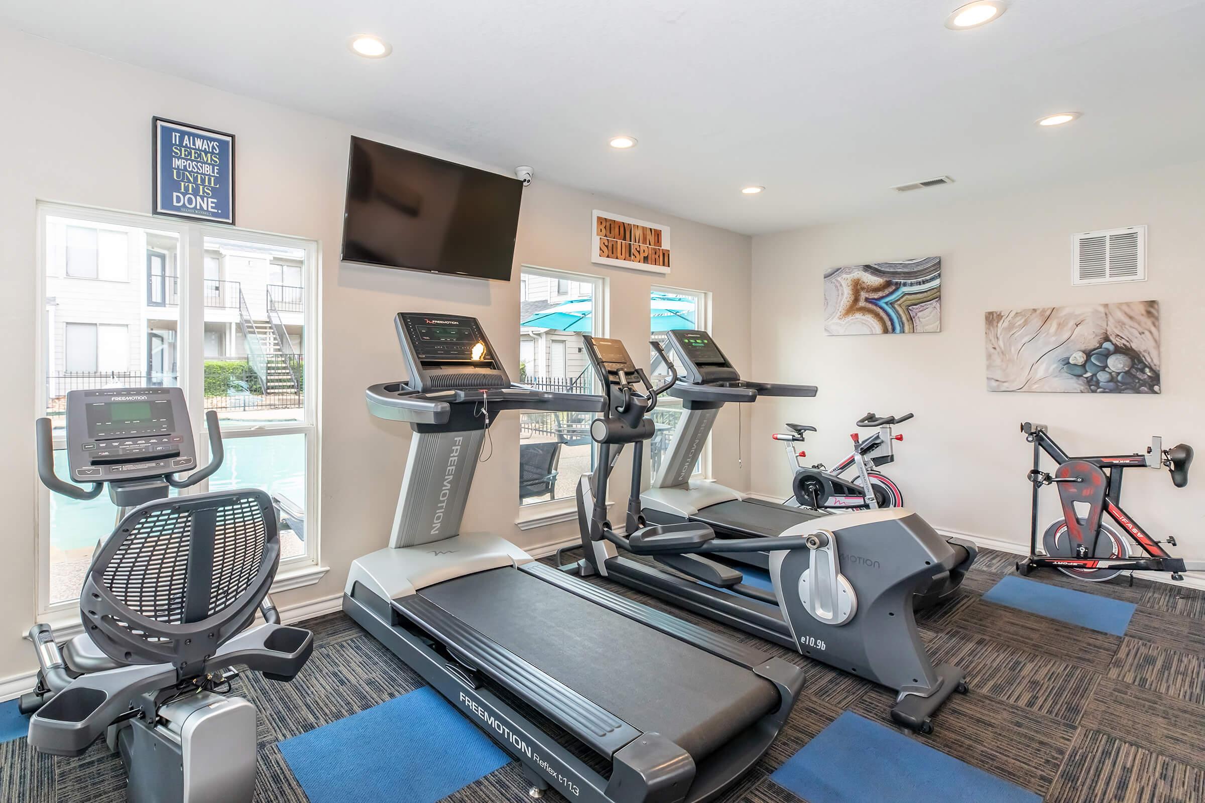 A modern gym featuring multiple exercise machines, including treadmills, a stationary bike, and a rowing machine. Large windows allow natural light to fill the space, and motivational wall art is displayed. The area is well-maintained, with blue mats on the floor and a view of a pool outside.