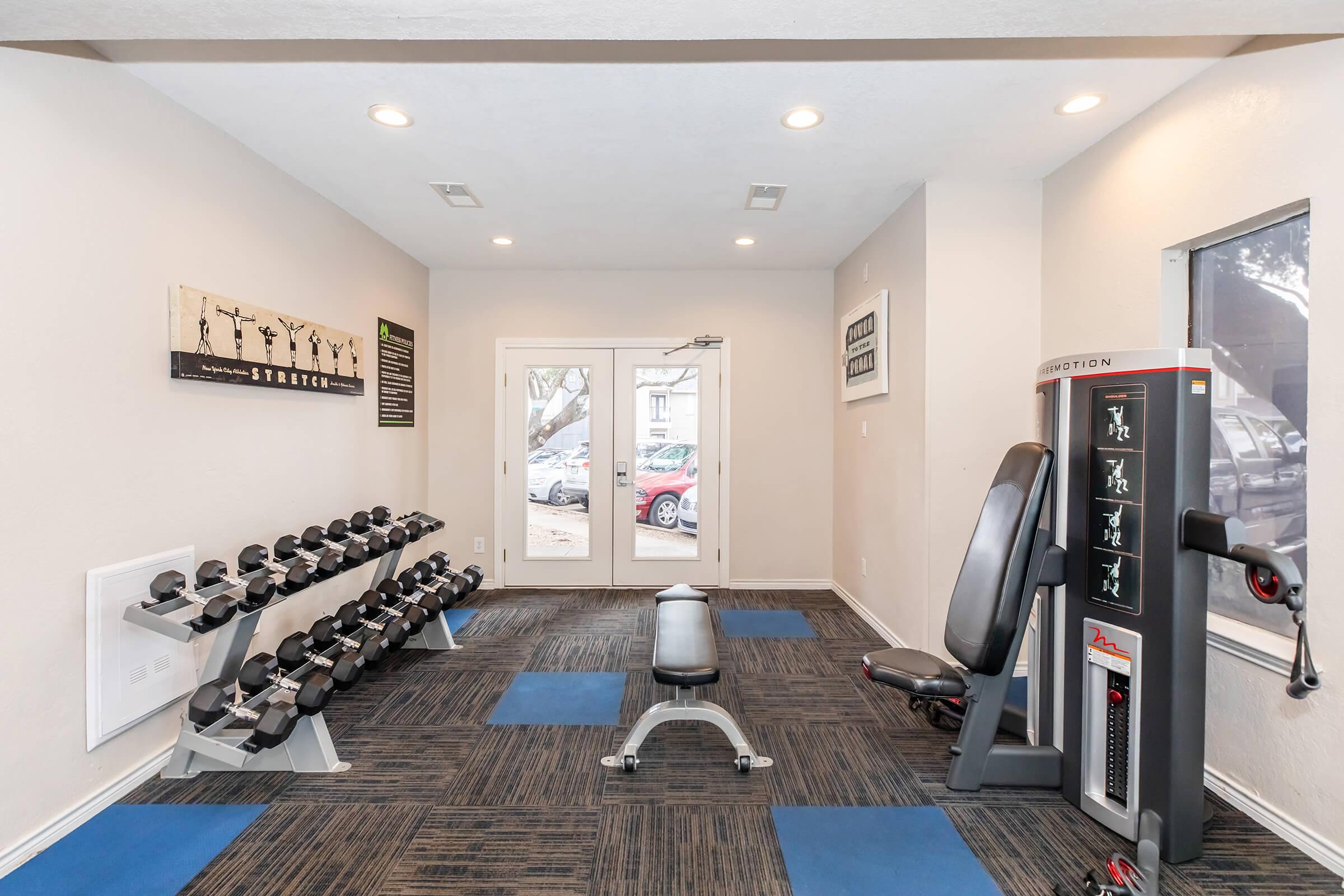 A modern fitness room featuring a set of dumbbells on a rack, a padded workout bench, and a multi-gym machine. The space has neutral-colored walls and blue carpet tiles, with large windows allowing natural light and a view of parked cars outside. Motivational wall art is visible.