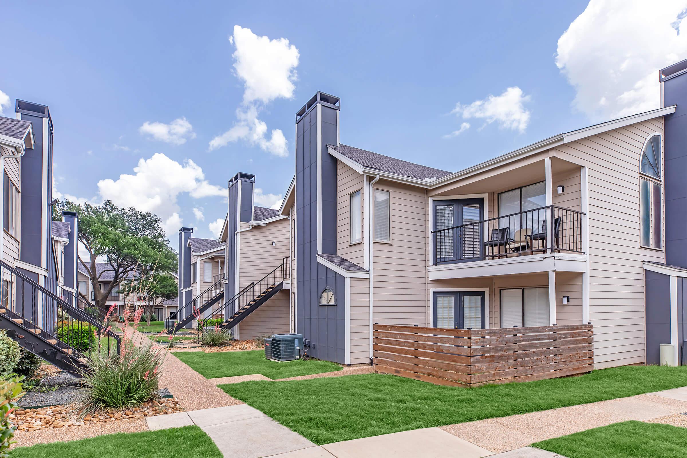 Two-story apartment buildings with balconies, surrounded by well-maintained lawns and landscaped gardens. Pathways lead to each unit, and there are outdoor features like wooden fences and decorative plants. The sky is partly cloudy, creating a bright atmosphere.