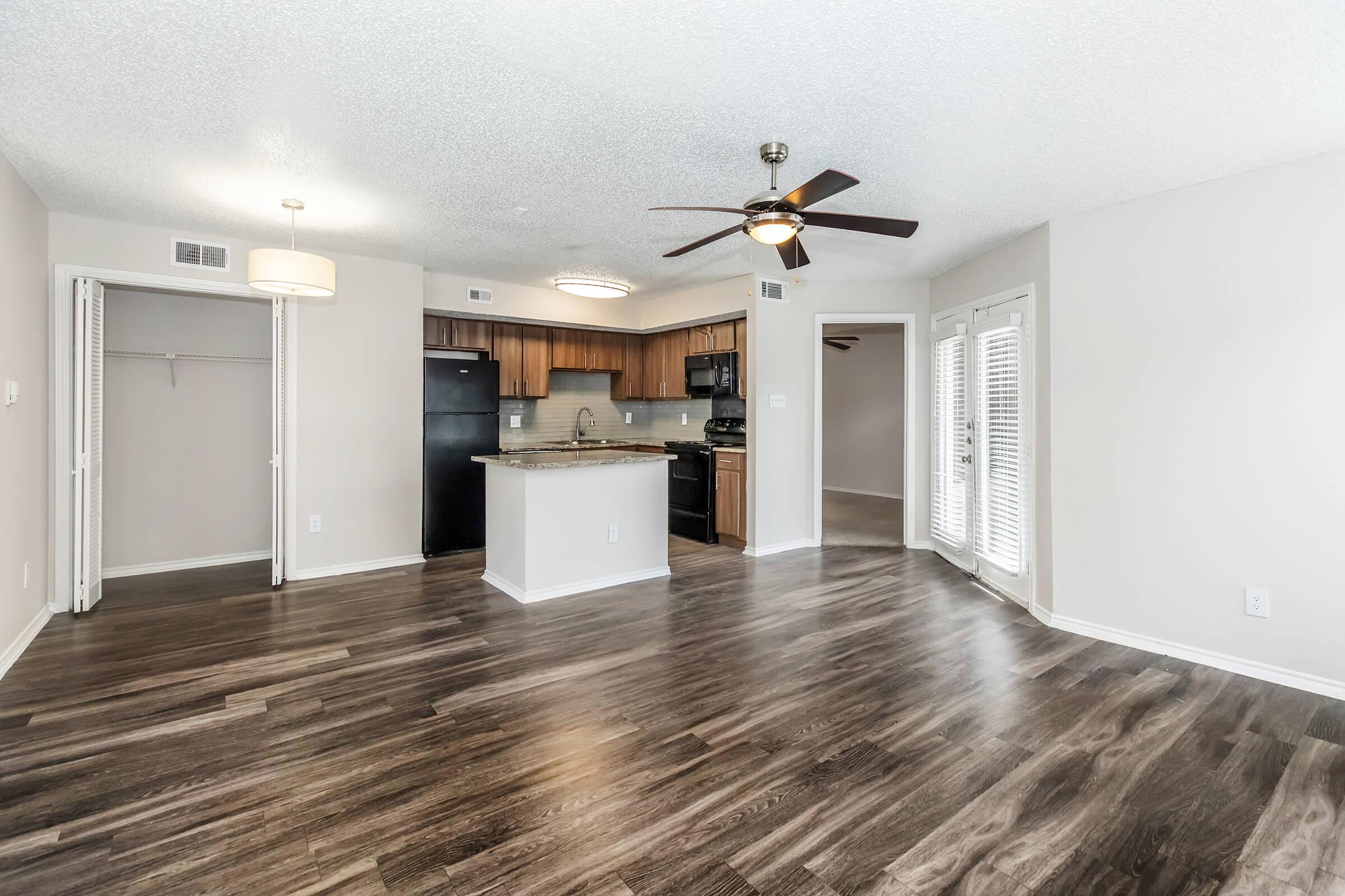 Spacious living area featuring modern hardwood flooring, a ceiling fan, and recessed lighting. An open kitchen with dark cabinets and appliances, and large windows allowing natural light. Includes a closet and access to a balcony or patio area through sliding doors.