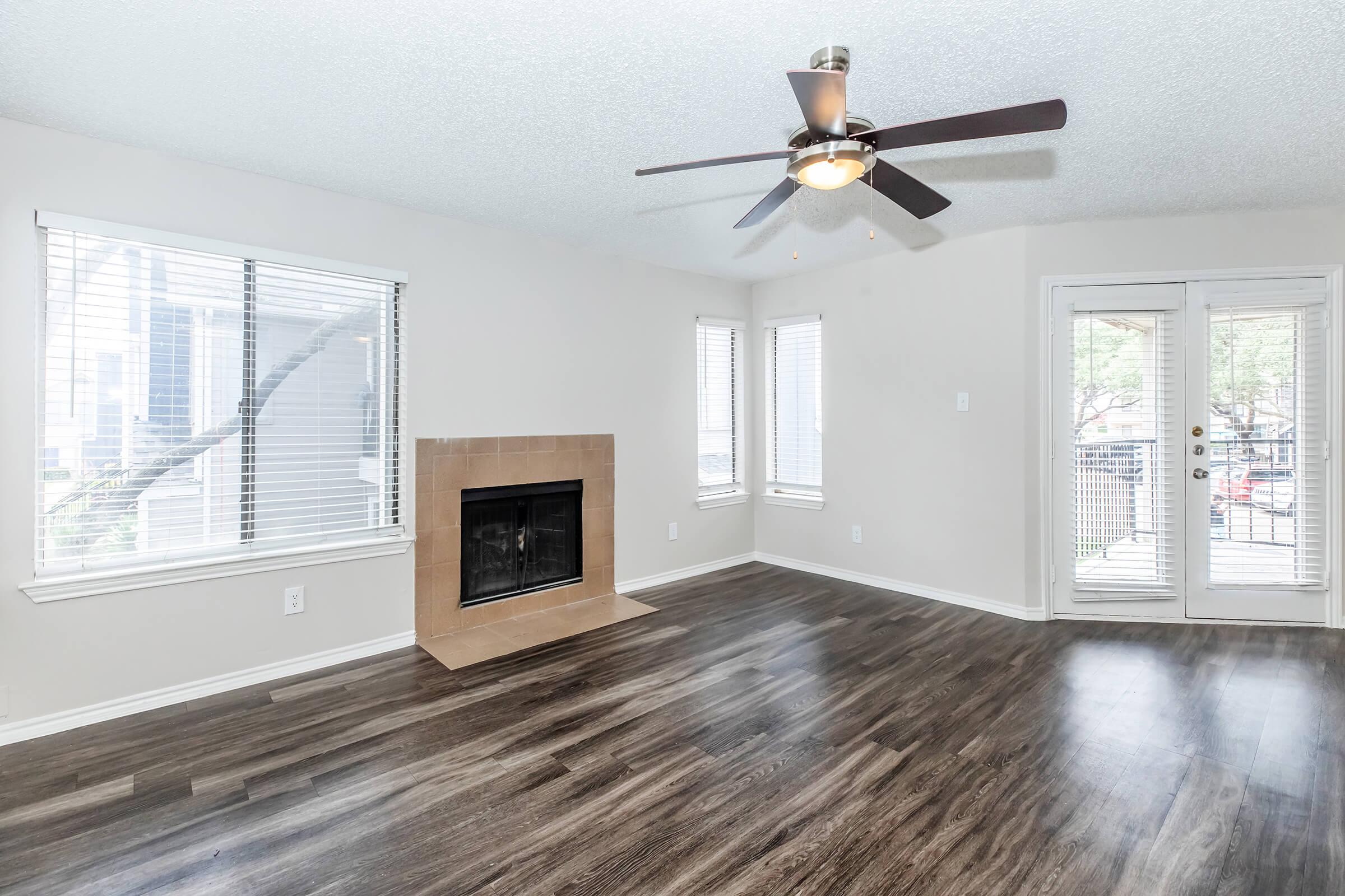A spacious living room featuring a ceiling fan, a fireplace, and large windows with blinds. The floor is covered with dark wood laminate, and there are doors leading to an outdoor area. The walls are painted in a light color, creating a bright and inviting atmosphere.