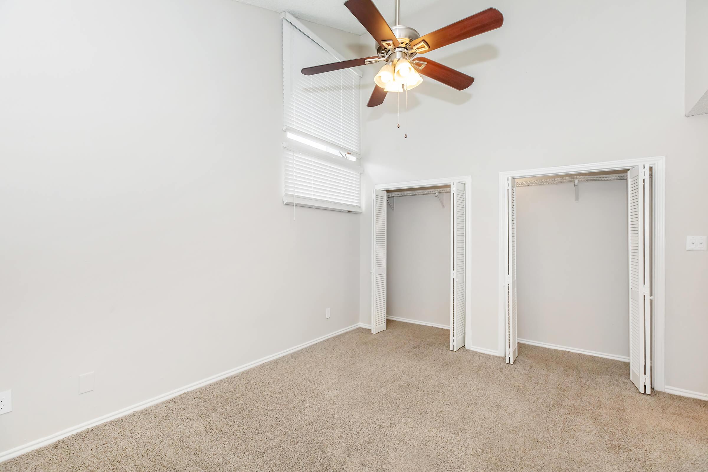 A well-lit, empty bedroom featuring beige carpet, a ceiling fan with wooden blades, and two white double-door closets. The walls are light-colored, and there are tall windows with blinds letting in natural light.